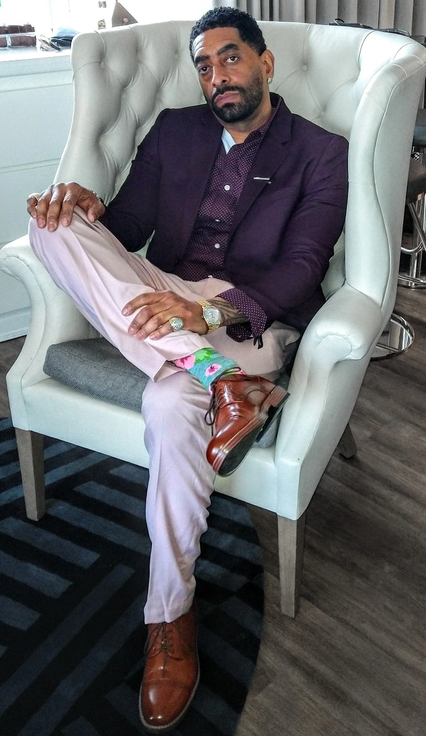 A man dressed in a dark blazer, dress shirt, and beige trousers, sitting on a white high-backed chair with a tufted design. He has dark curly hair, a beard, and is wearing jewelry, including rings and watches. His right leg is crossed over his left knee, showing colorful floral socks and brown leather shoes. He has a serious or contemplative expression.