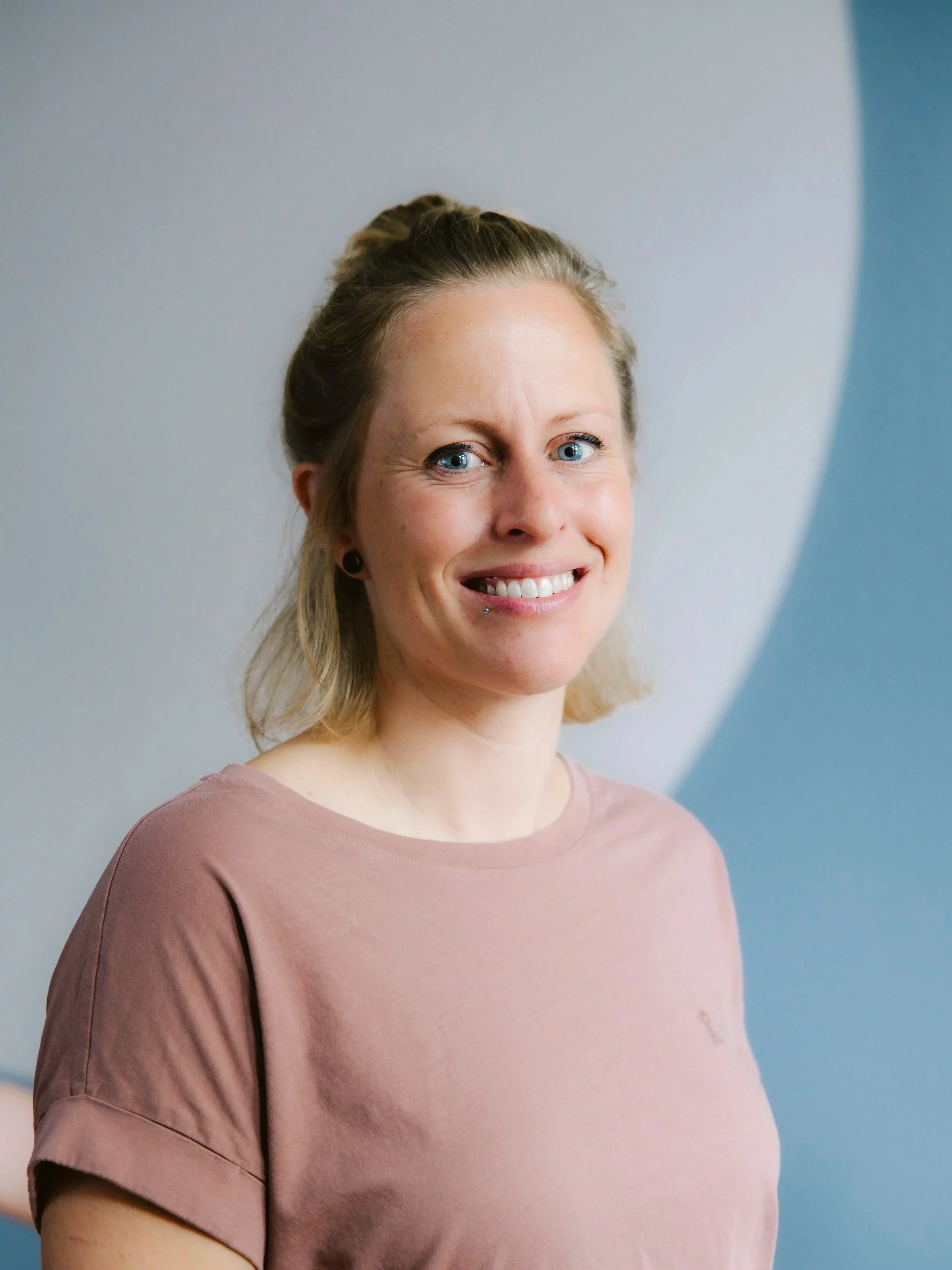 Portrait von Manuel Wiedmer eine Frau mit blauen Augen, hellblonden Haaren, die nach hinten hochgesteckt sind, trägt ein rosa T-Shirt, lächelt und steht vor einer blauen und weißen Wand.