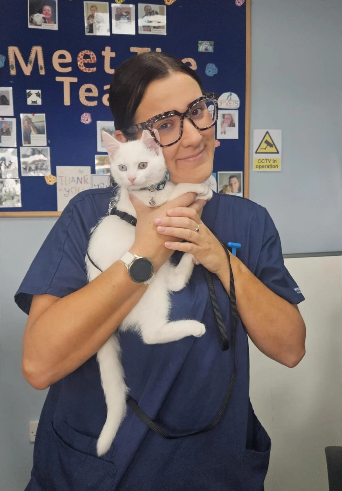 A woman wearing glasses and a navy uniform holds a white cat with blue eyes in front of a bulletin board that says 'Meet the Team'.