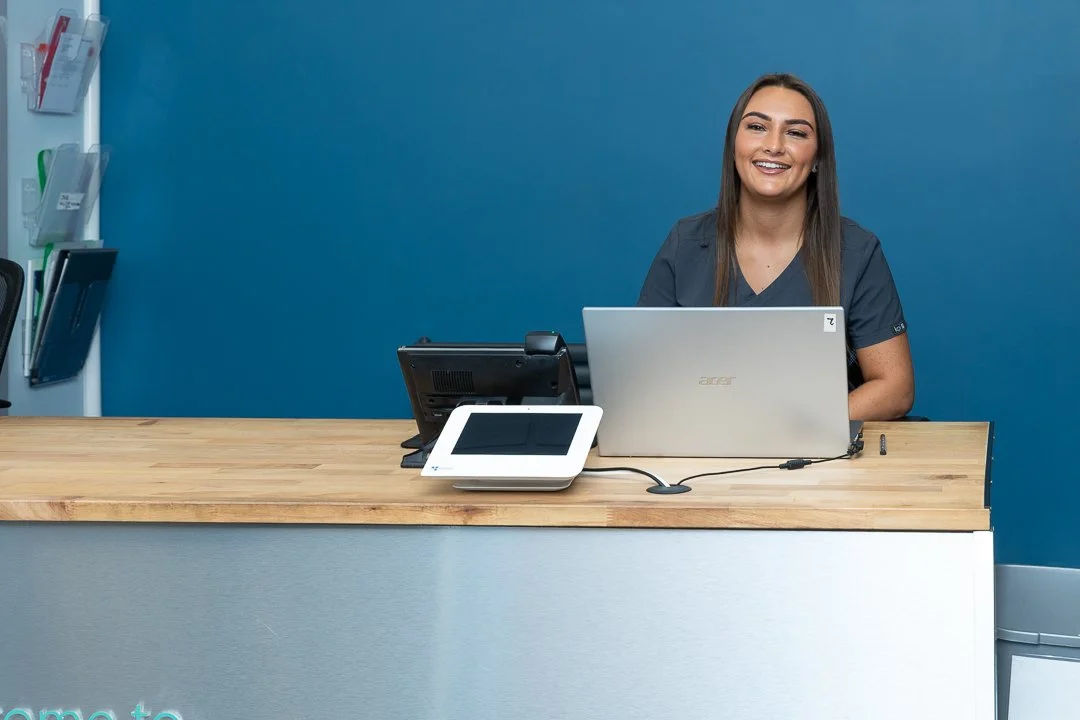 A smiling woman wearing dark scrubs sitting at a reception desk with a laptop, a tablet, and a phone in a modern office or clinic setting.