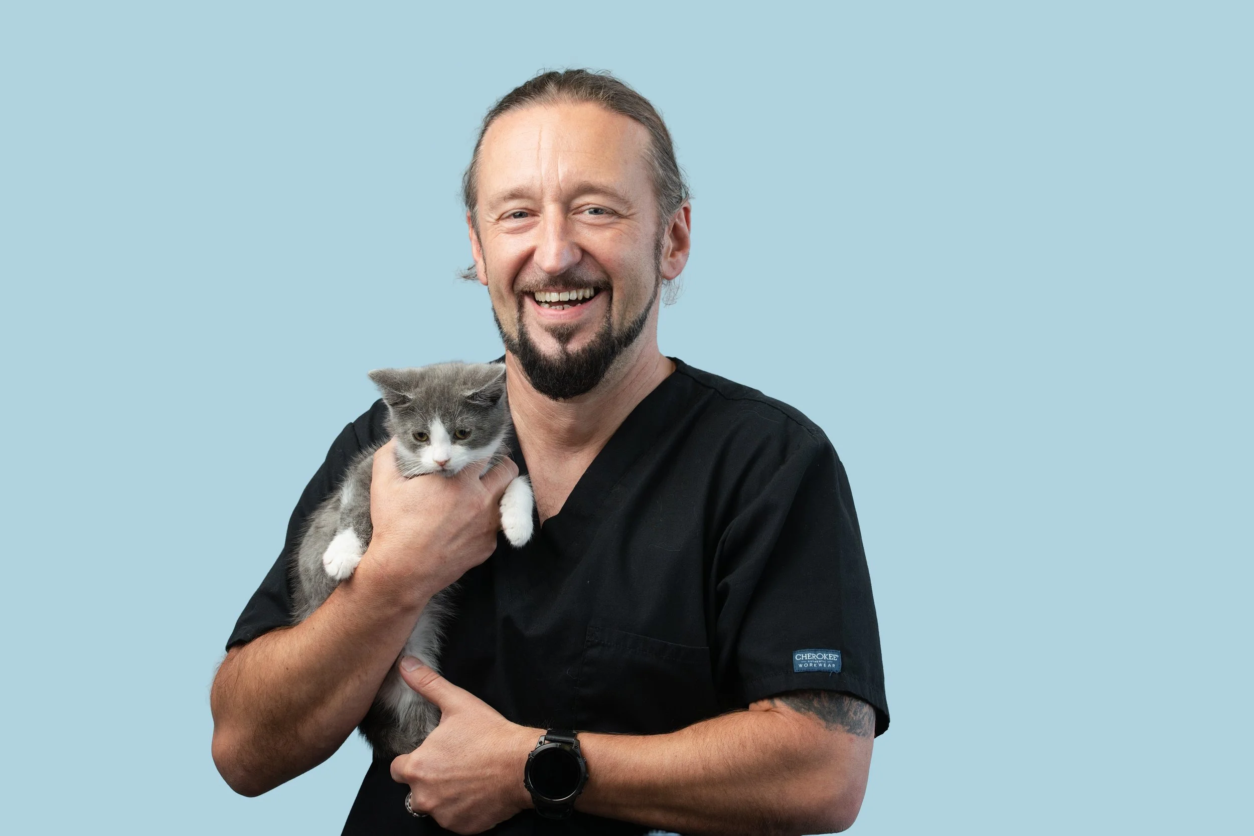 A man with a beard and long hair holding a gray and white kitten, smiling against a light blue background.