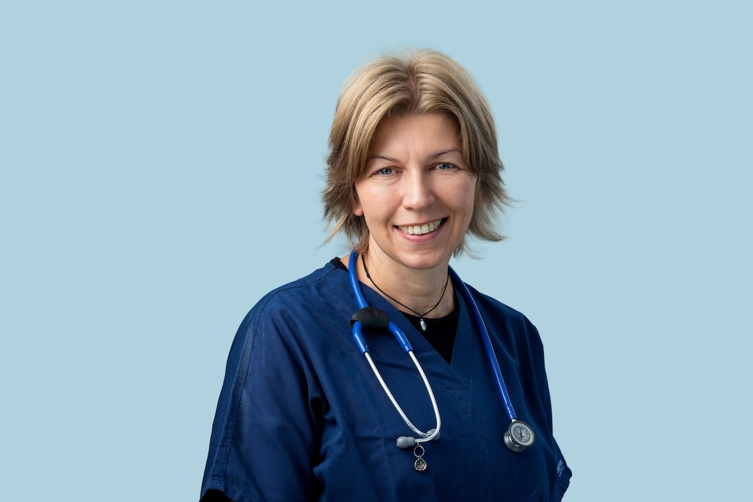 A veterinarian wearing a dark uniform, named Dominika, examines a newborn puppy on a yellow towel on an examination table in a veterinary clinic.