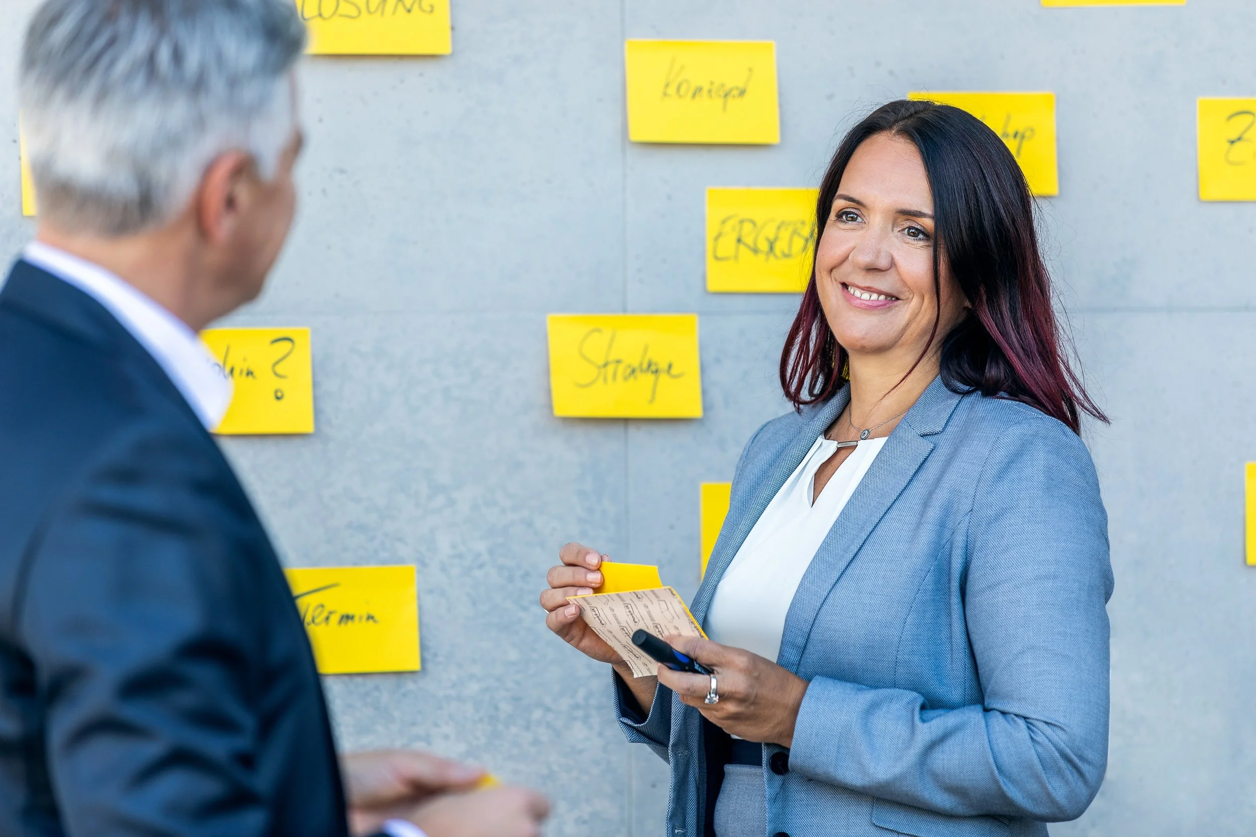 Zwei Berufstätige, eine Frau und ein Mann, im Gespräch vor einer Wand mit gelben Post-its. Die Frau hat dunkle Haare mit Rot-Tupfen und trägt einen grauen Blazer, während der Mann eine dunkle Jacke trägt.
