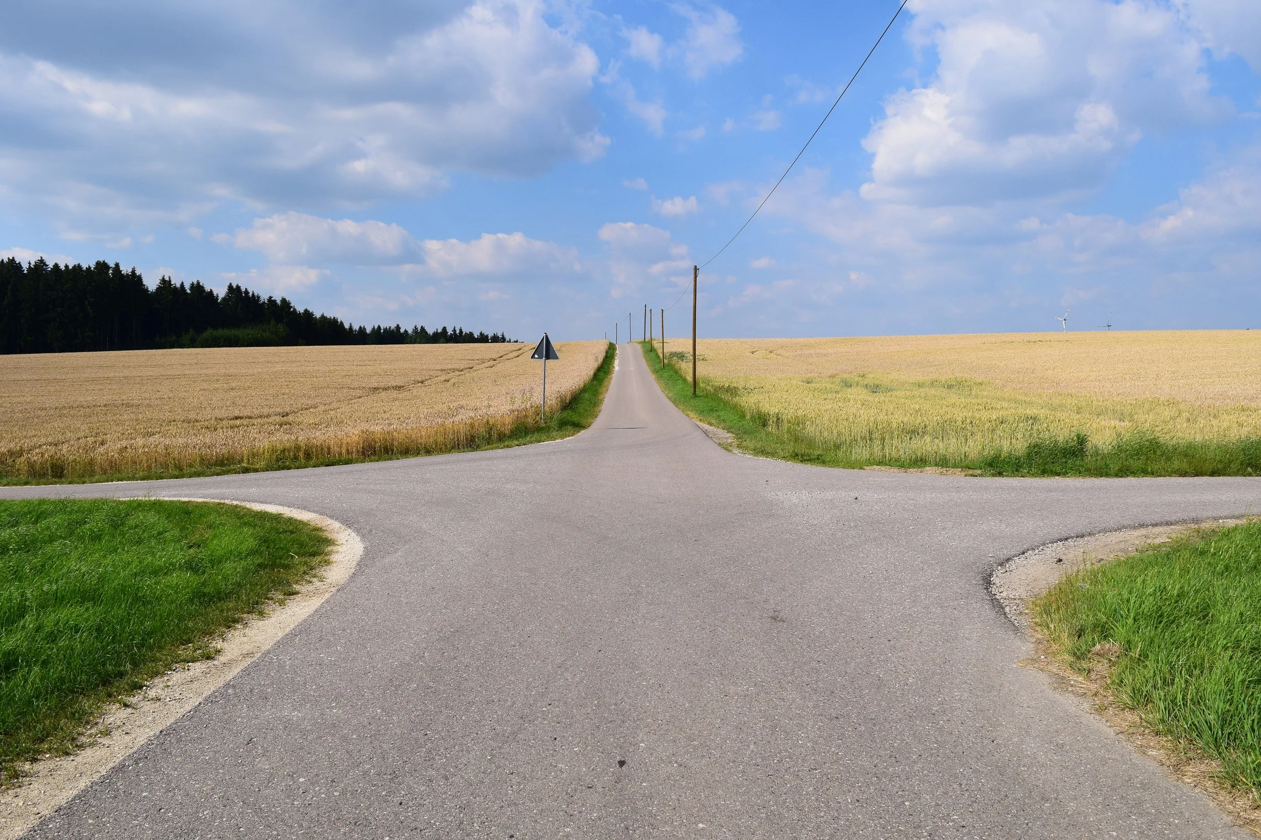 Eine landwirtschaftliche Straße, die sich teilt und in die Ferne führt, umgeben von Getreidefeldern bei sonnigem Himmel mit Wolken.