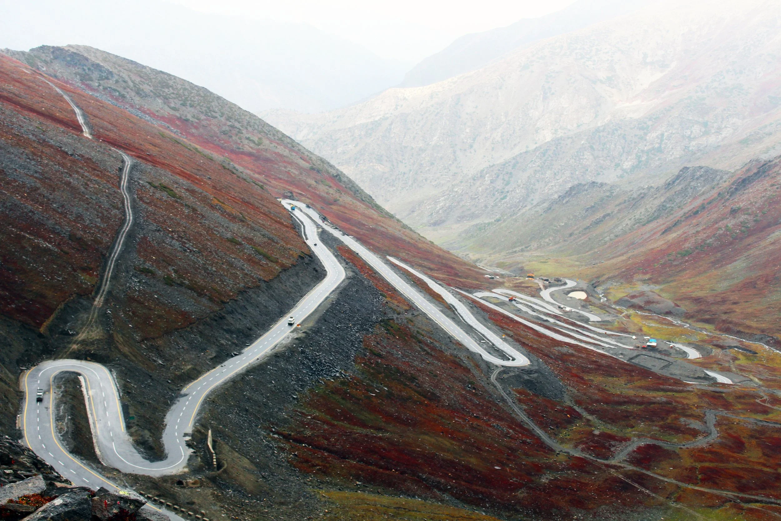 Serpentine Straße windet sich durch bergiges, herbstliches Gelände mit roten und braunen Vegetation. Nebel und Berge im Hintergrund.