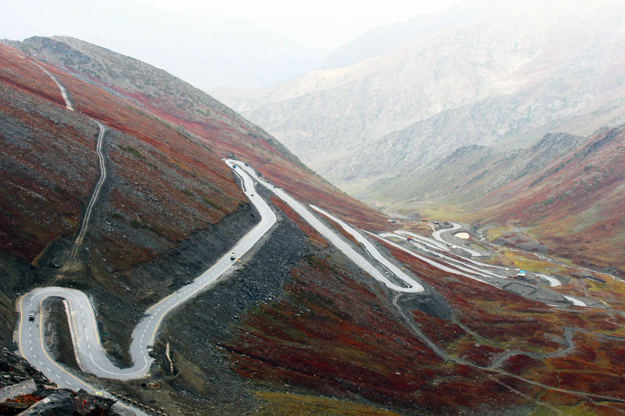 Schleifenförmige Straße windet sich durch eine bergige, herbstliche Landschaft mit rot und braun gefärbtem Laub und steil abfallenden Hängen.