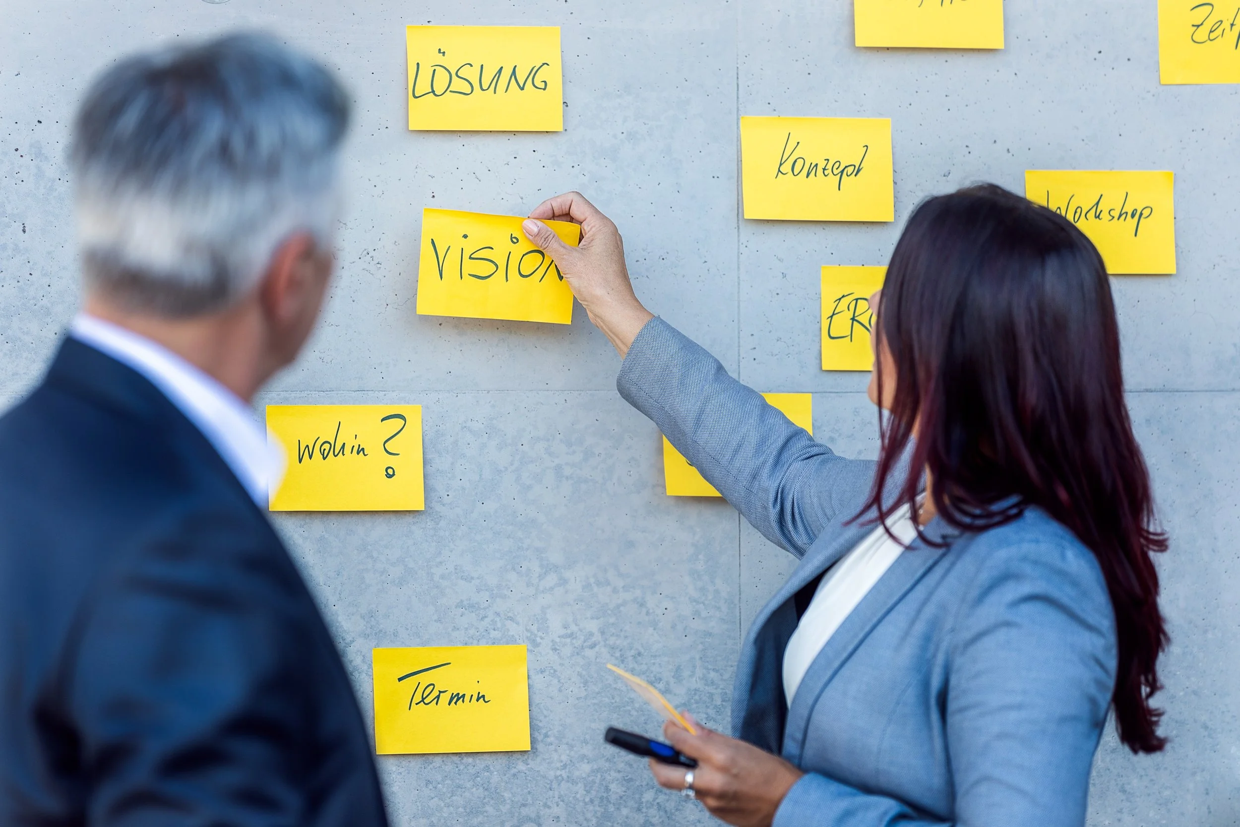 Zwei Geschäftsleute vor einer Wand mit gelben Haftnotizen, auf denen Wörter wie 'Vision', 'Lösung', 'Konzept' und 'Termin' geschrieben sind.