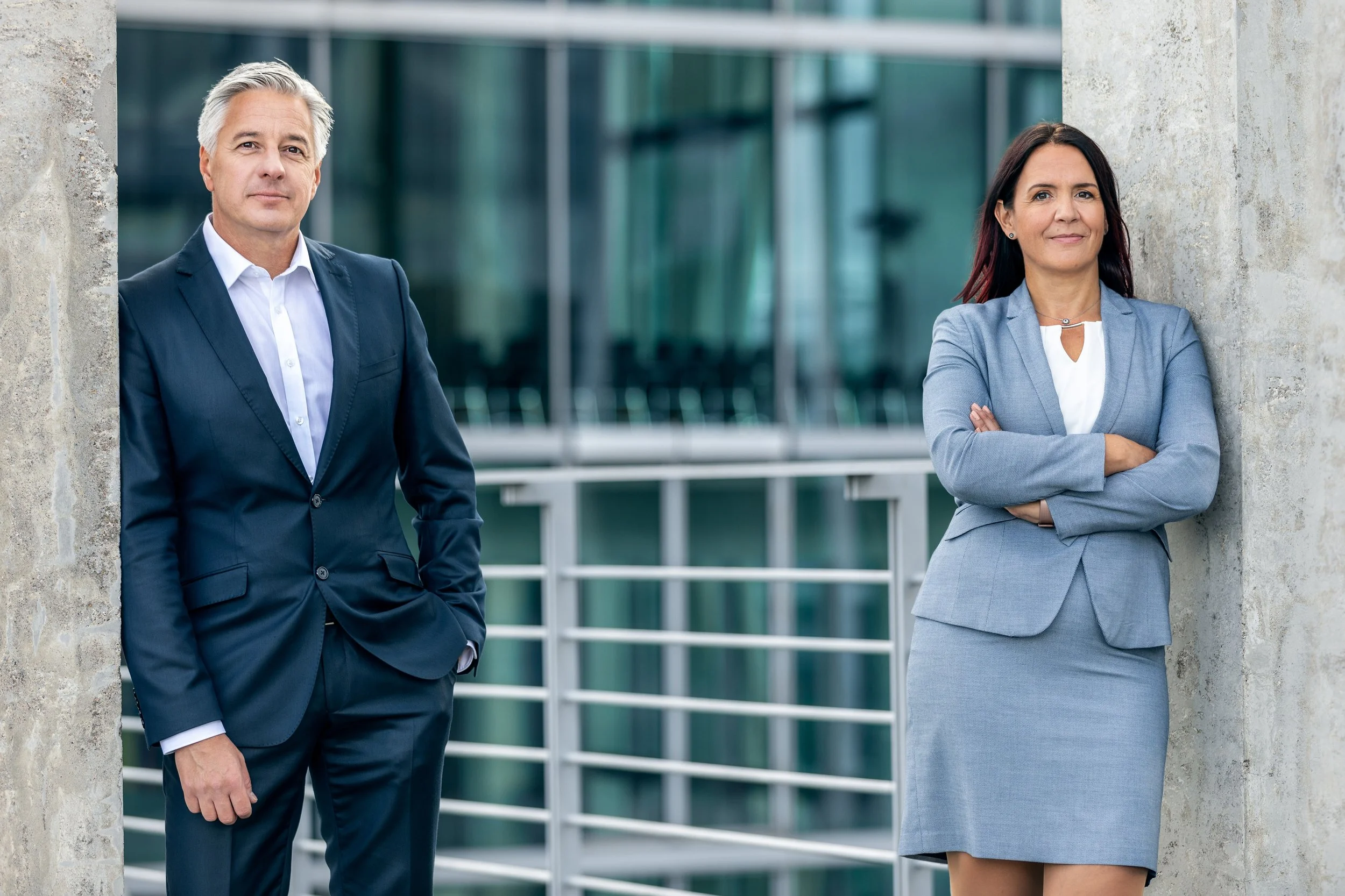 Zwei berufstätige Personen, ein Mann und eine Frau, posieren nebeneinander vor einem modernen Bürogebäude. Der Mann trägt einen dunklen Anzug mit weißem Hemd, die Frau trägt einen grauen Businessanzug mit weißem Top. Beide schauen selbstbewusst in die Kamera.