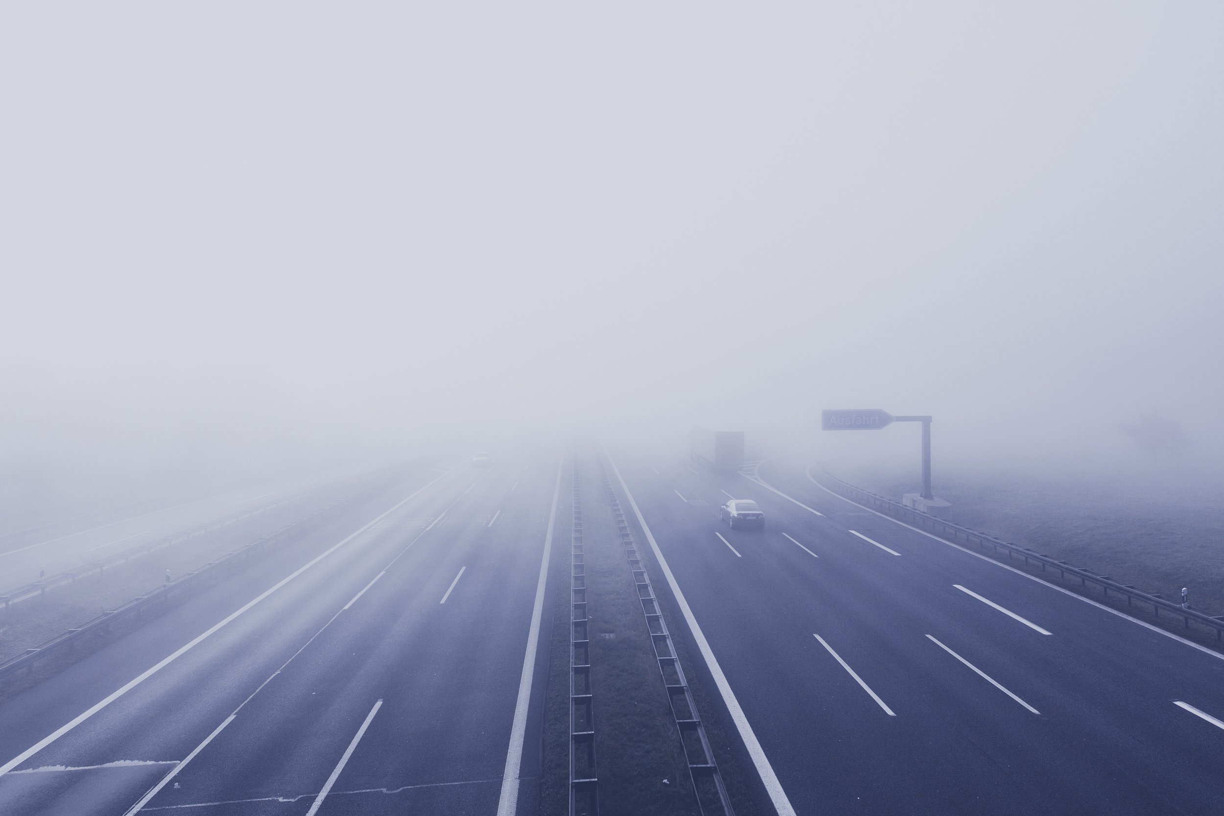Leerer Autobahnabschnitt in dichtem Nebel, wenige Autos sichtbar, Verkehrsschilder im Hintergrund.