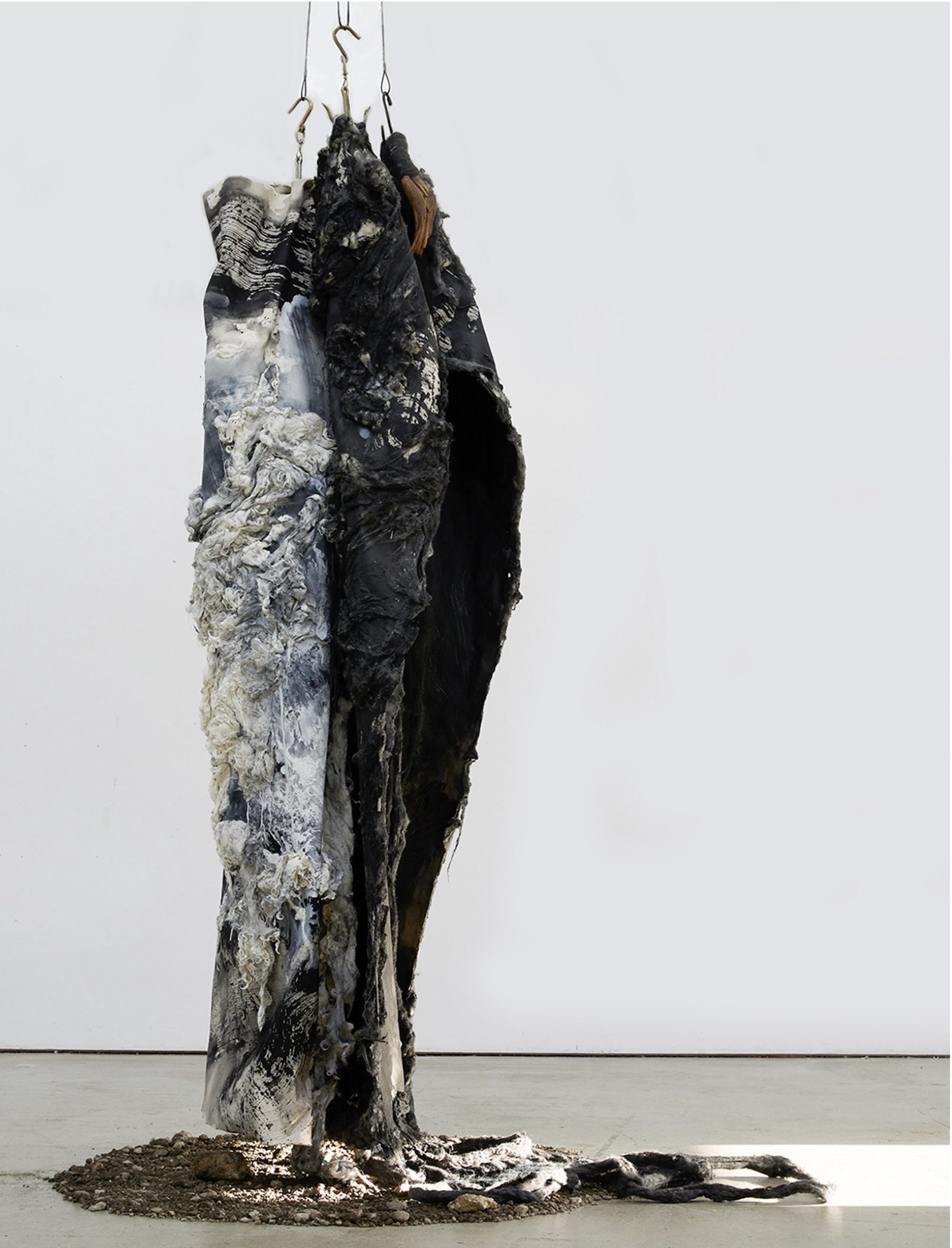 Tall hanging sculpture by Monica Perez made of charred, wool-like and clay textures, suspended above a mound of earth on the gallery floor.