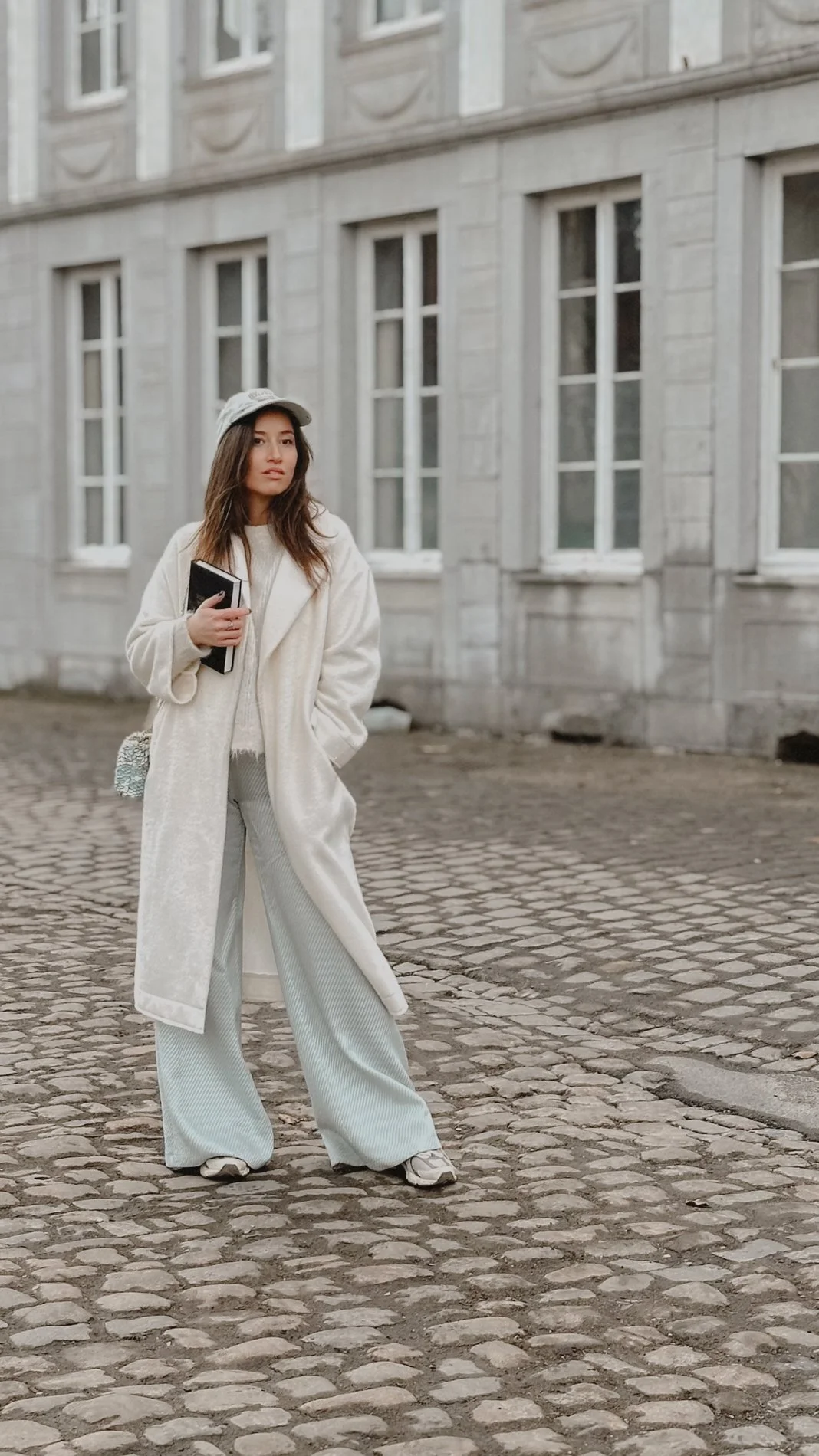 Young woman wearing a white coat, cream sweater, light-colored wide-leg pants, sneakers, and a cap, standing on cobblestone street in front of a gray stone building with multiple windows. She holds a notebook in her right hand.