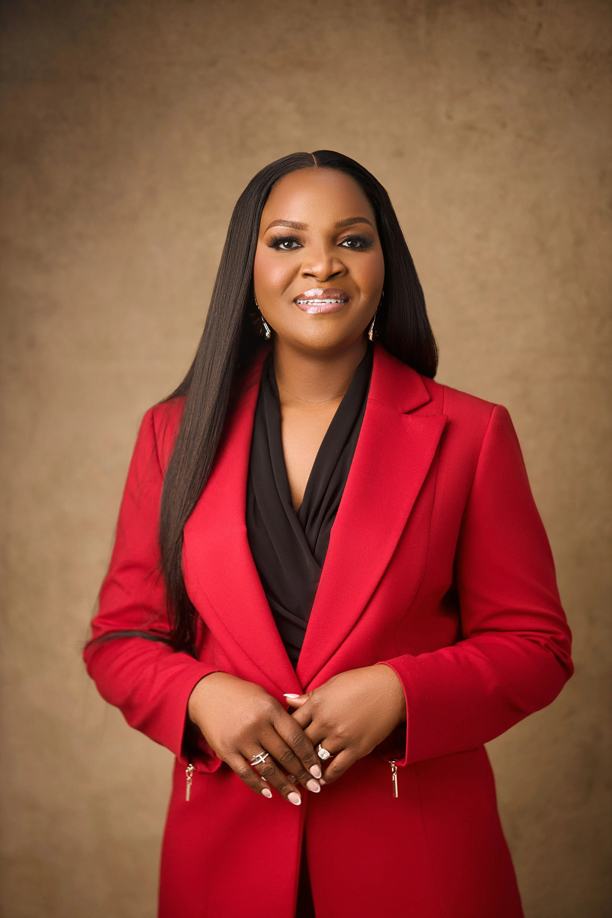 A woman with dark hair and makeup, wearing a red blazer with a black blouse, standing against a gold background.