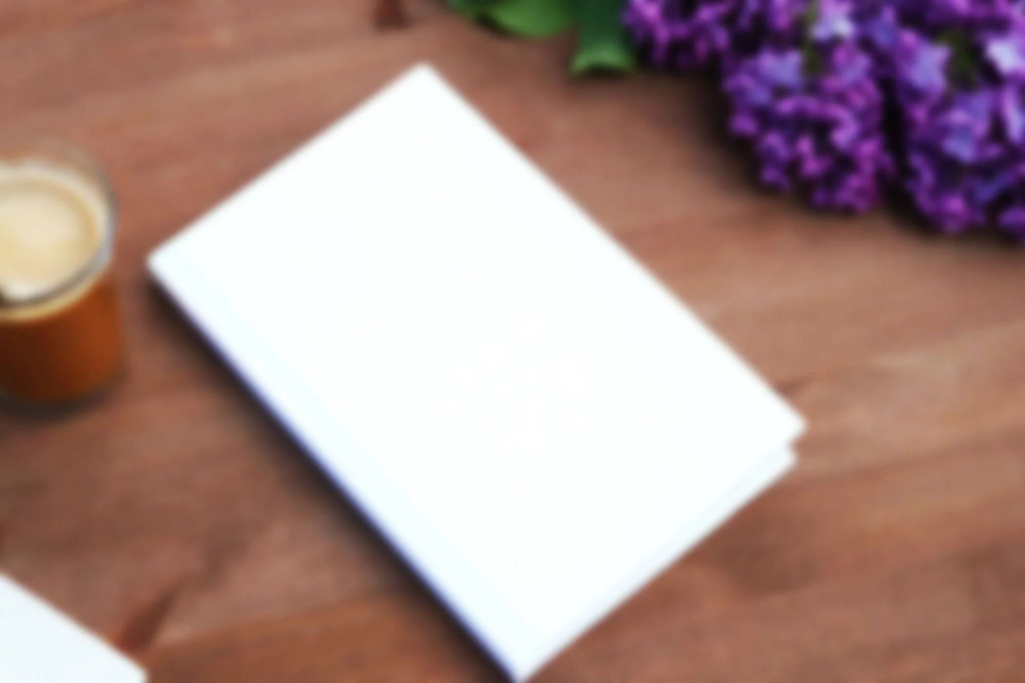 A white notebook on a wooden table with a glass of coffee on the left and purple flowers in the upper right corner.