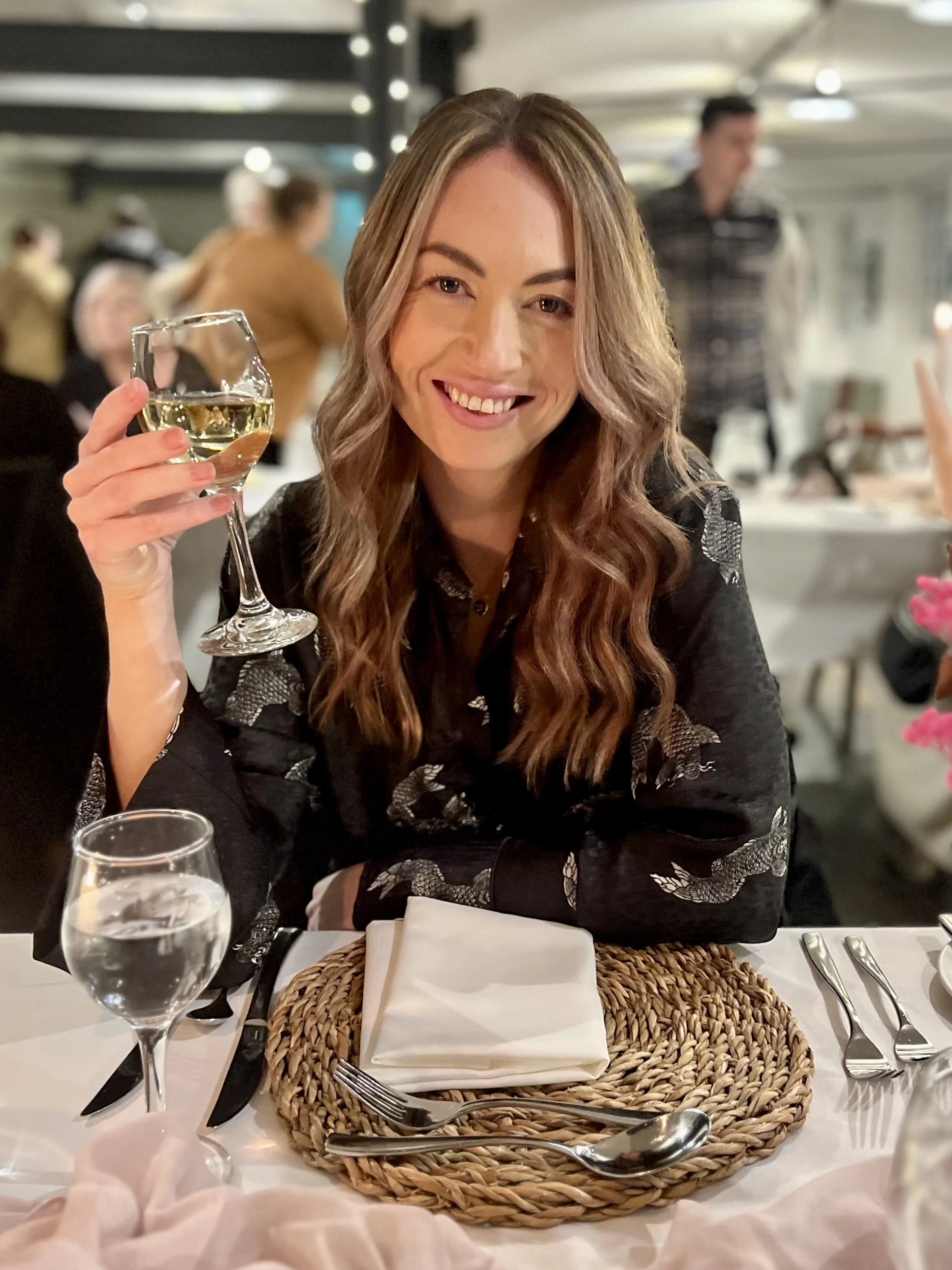 A woman with wavy brown hair smiling and holding a glass of white wine at a dinner table in a restaurant.