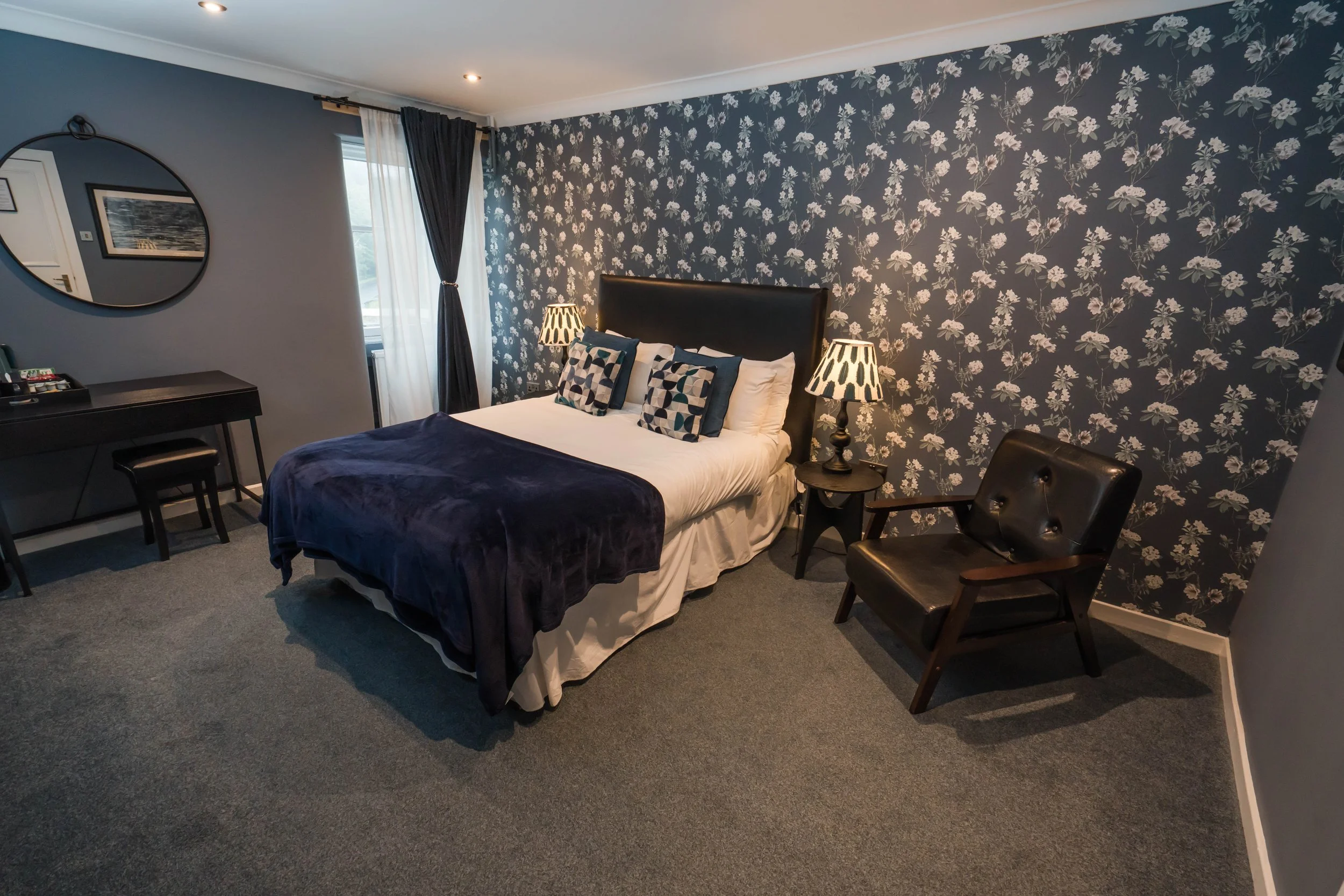A neatly made bed with white and black pillows in a modern hotel room, flanked by mirrored nightstands with black lamps, next to a window with black curtains and sheer white curtains.