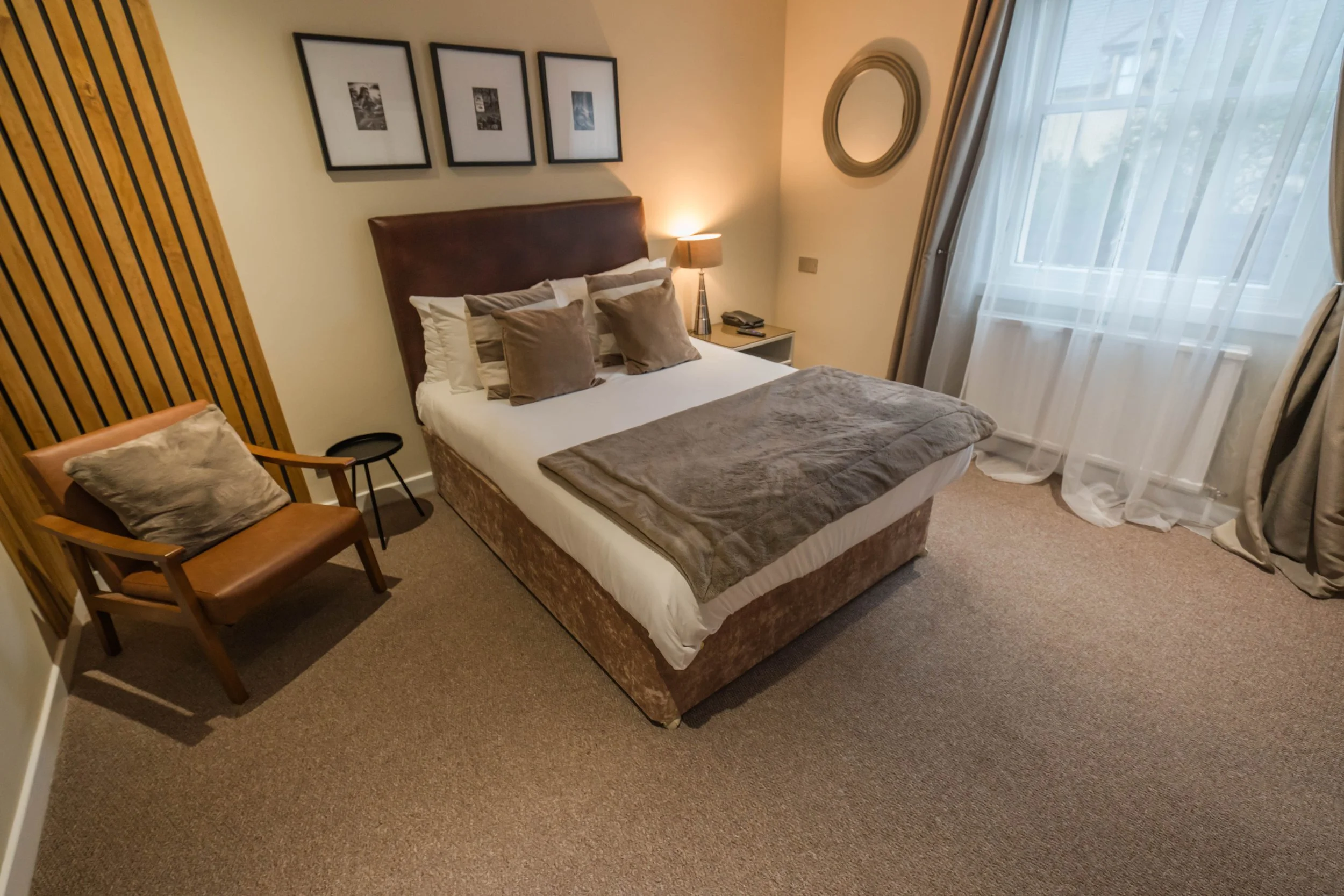 A cozy hotel room with a bed covered with a brown blanket, a window with sheer white curtains, a wall-mounted television, a small desk with a chair, a wardrobe, and decorative plants.