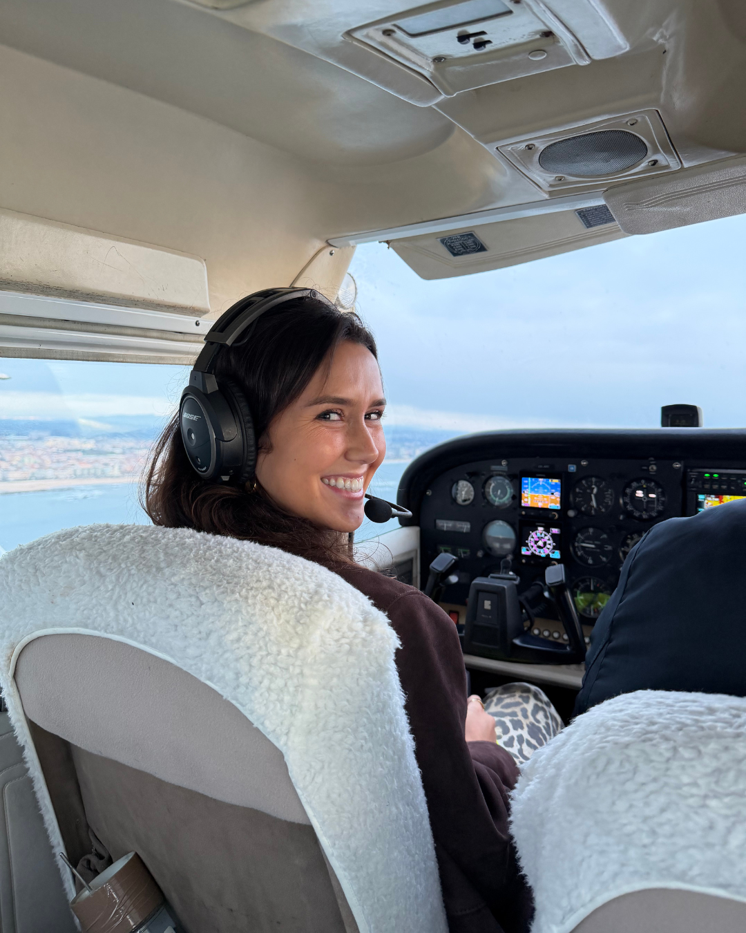Mulher sorrindo dentro do cockpit de um avião, usando fone de ouvido com microfone, com instrumentos de voo ao fundo e uma paisagem de cidade e rio visível pela janela.