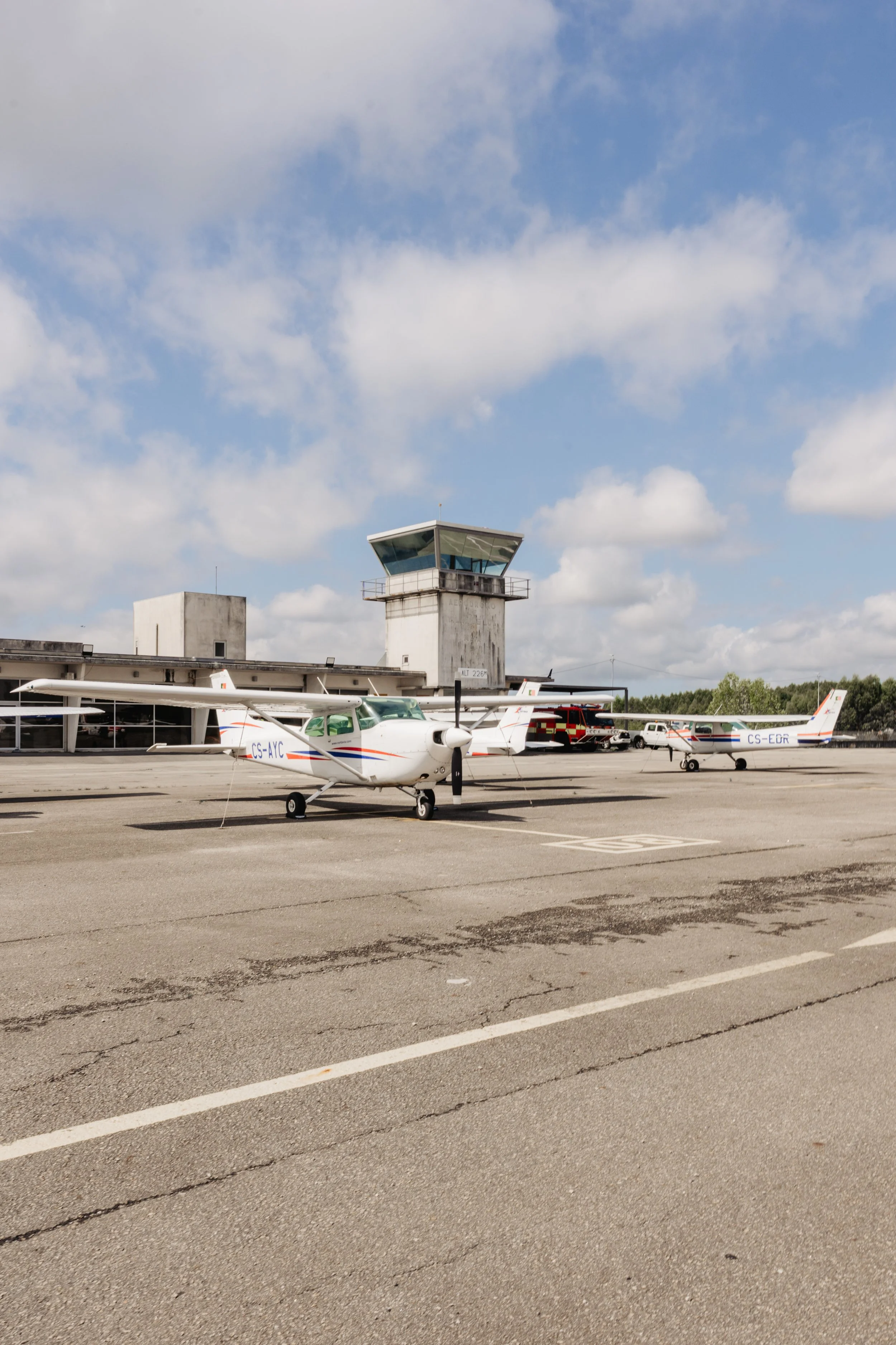 Aviões pequenos estacionados no aeroporto, com a torre de controle ao fundo, sob céu parcialmente nublado.