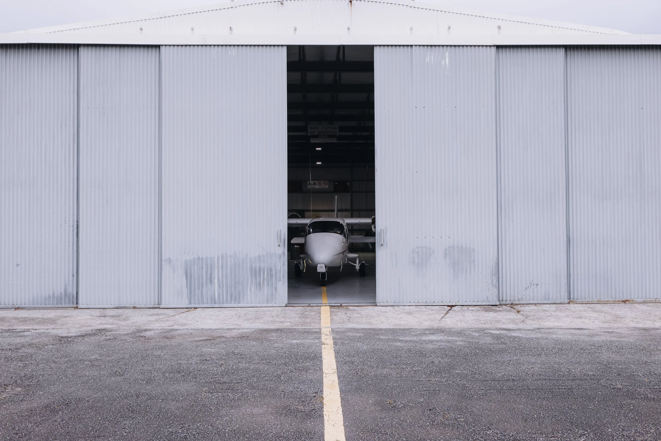 hangar com porta aberta, avião pequeno dentro dele, pista de estacionamento na frente