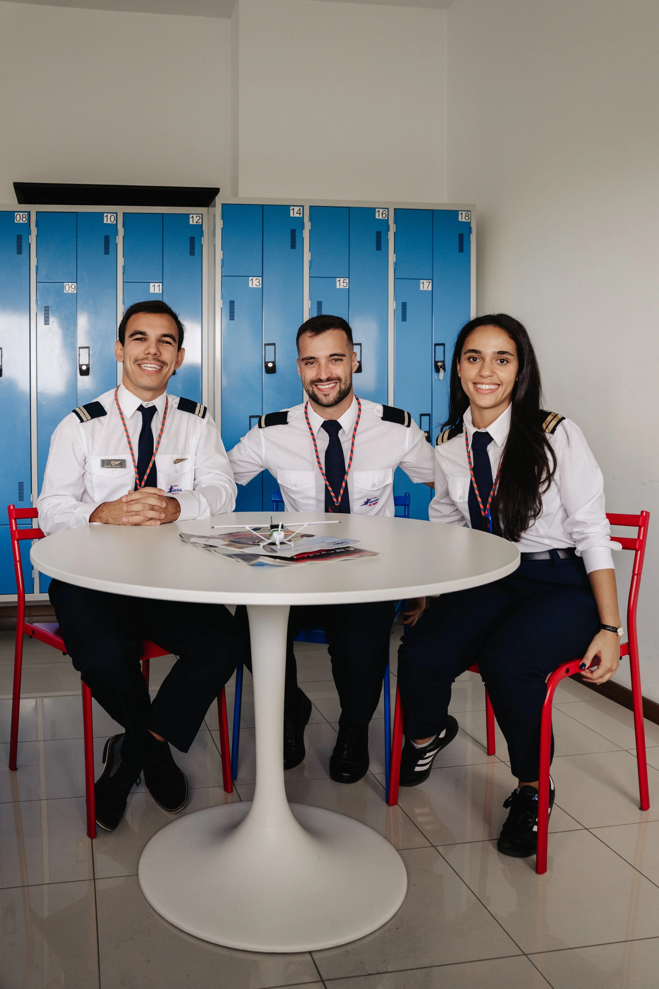 Três tripulantes de aviação sorridentes sentados em volta de uma mesa branca com lockers azuis ao fundo. Eles estão vestindo uniformes de pilotos.