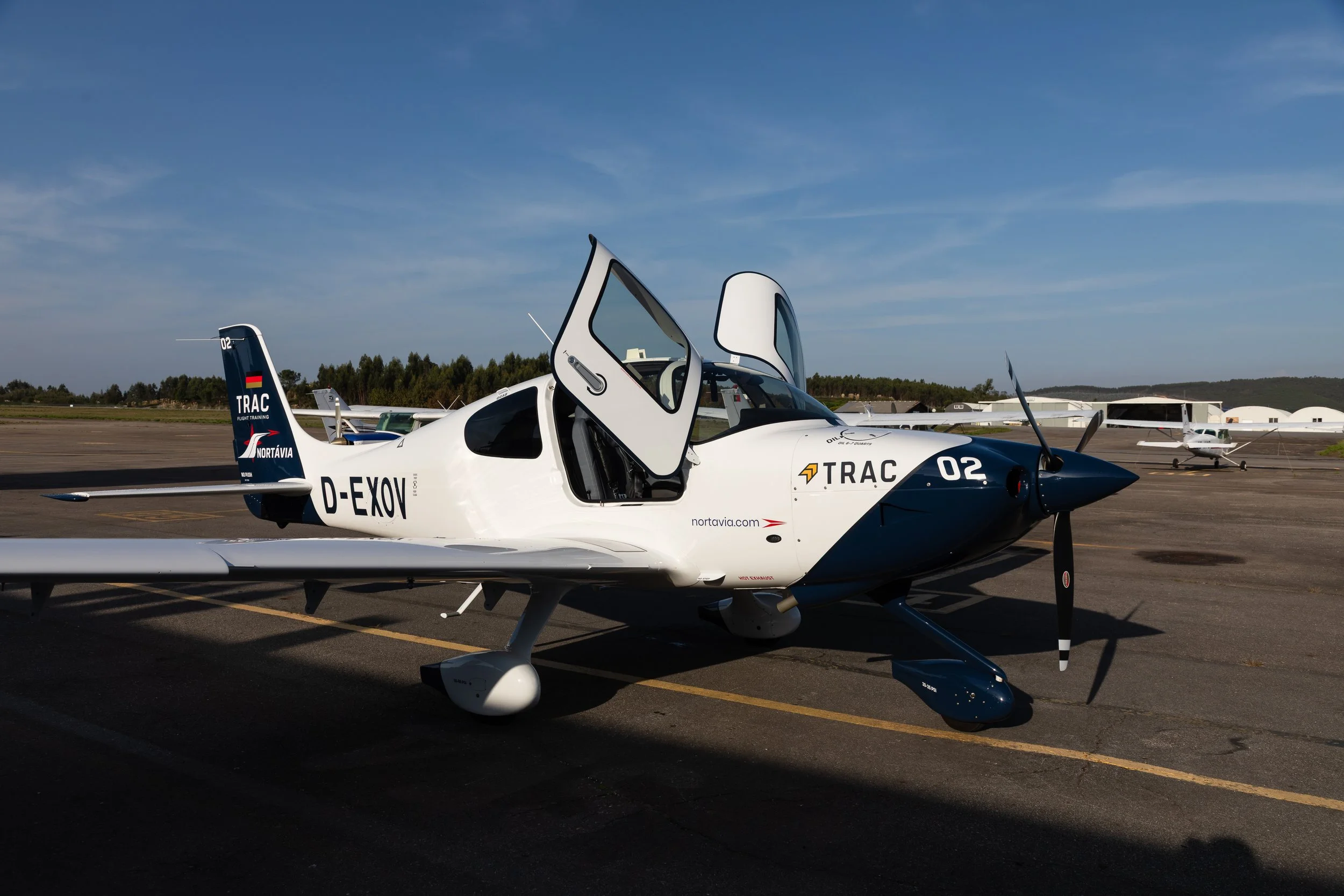 Avião de treino em solo de aeroporto, com o cockpit aberto, em primeiro plano, e outros aviões ao fundo, céu claro.