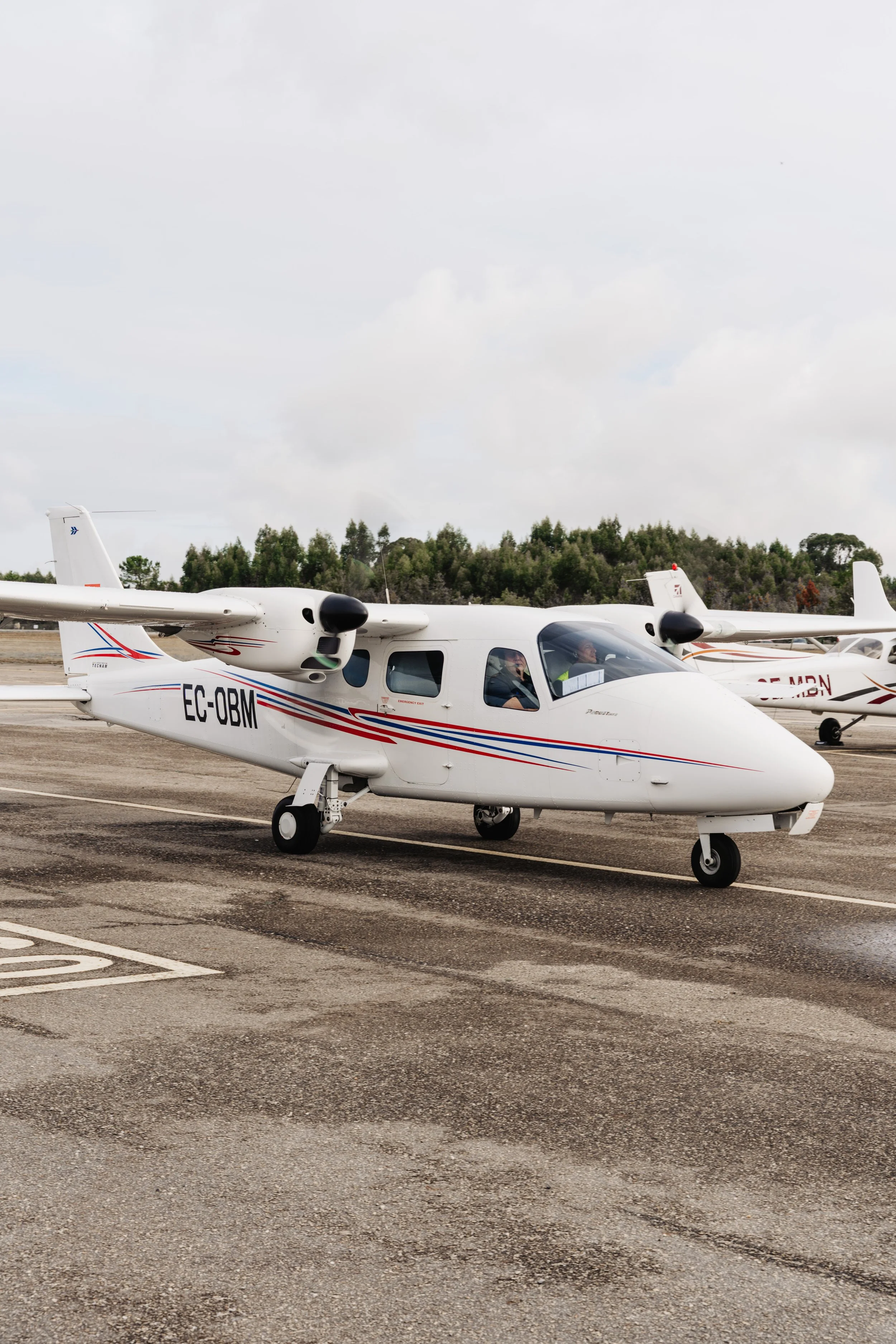 Avião pequeno de passageiros estacionado na pista do aeroporto com outros aviões ao fundo e árvores ao redor, sob céu nublado.