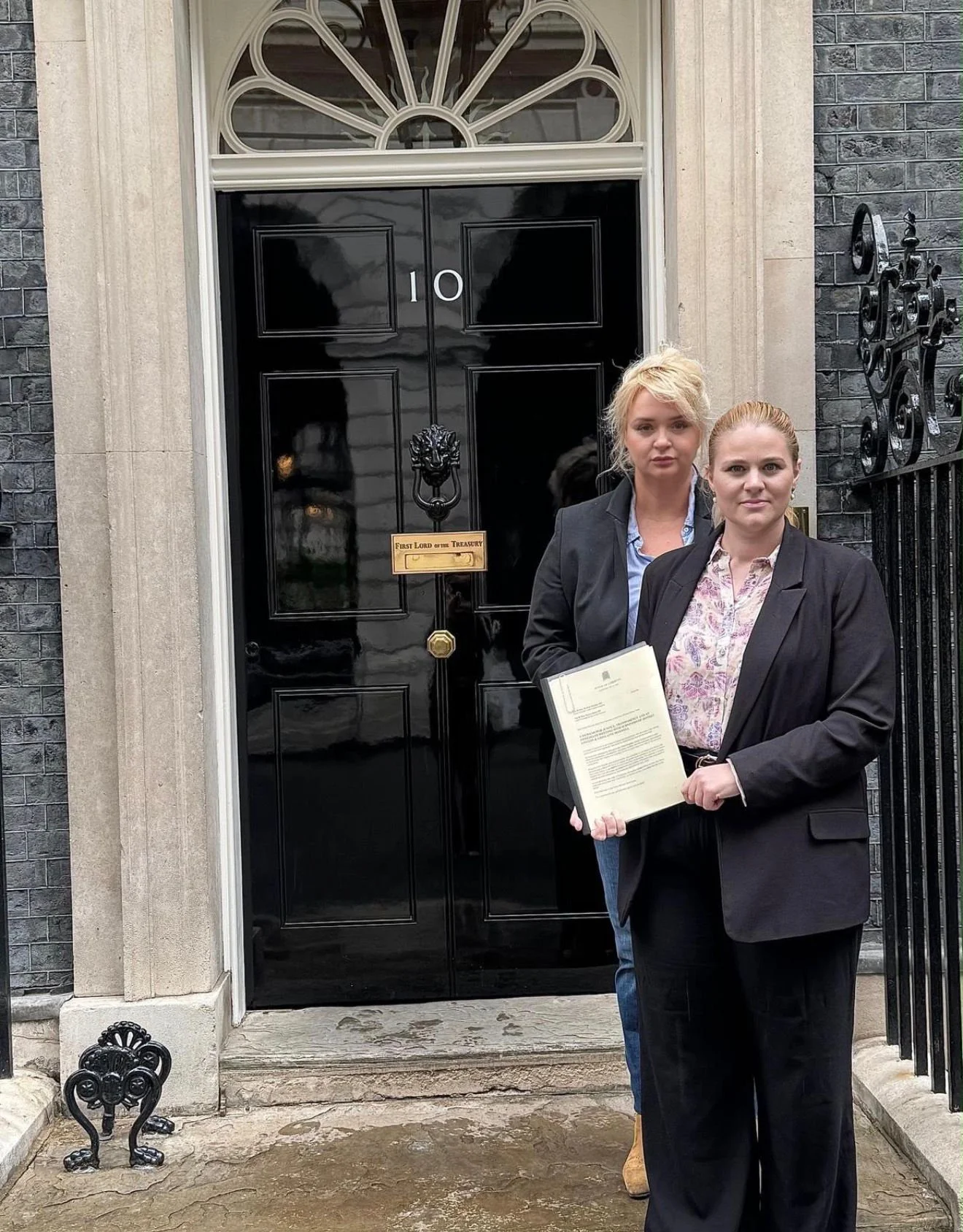 Two women standing in front of a black front door of a house with the number 10. The woman in the foreground is holding a document, wearing a black blazer, and has red hair. The woman behind her has blonde hair tied up, is dressed in a blazer and shirt, and is slightly smiling.