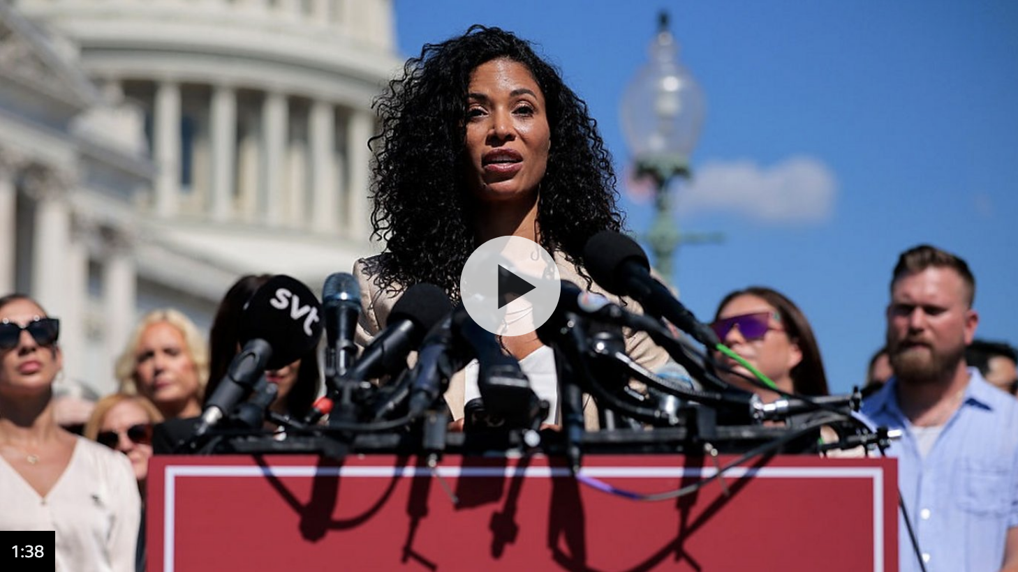 A woman with curly black hair speaking at a press conference with multiple microphones in front of her outdoors in front of the U.S. Capitol building. Several people are standing behind her.