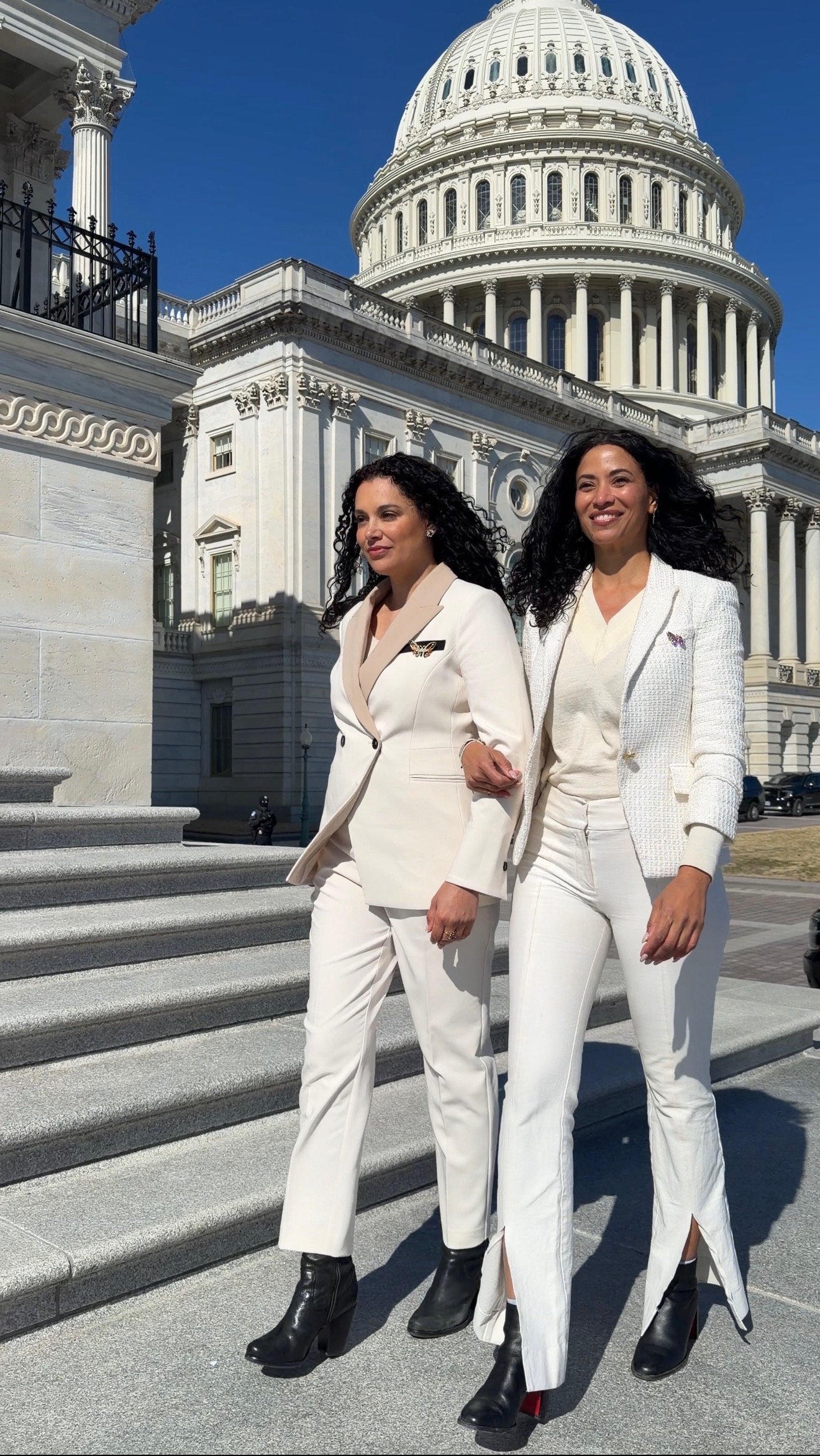 Two women in white outfits walking down the steps of a government building with a large dome, likely the U.S. Capitol. They are dressed professionally and are smiling.