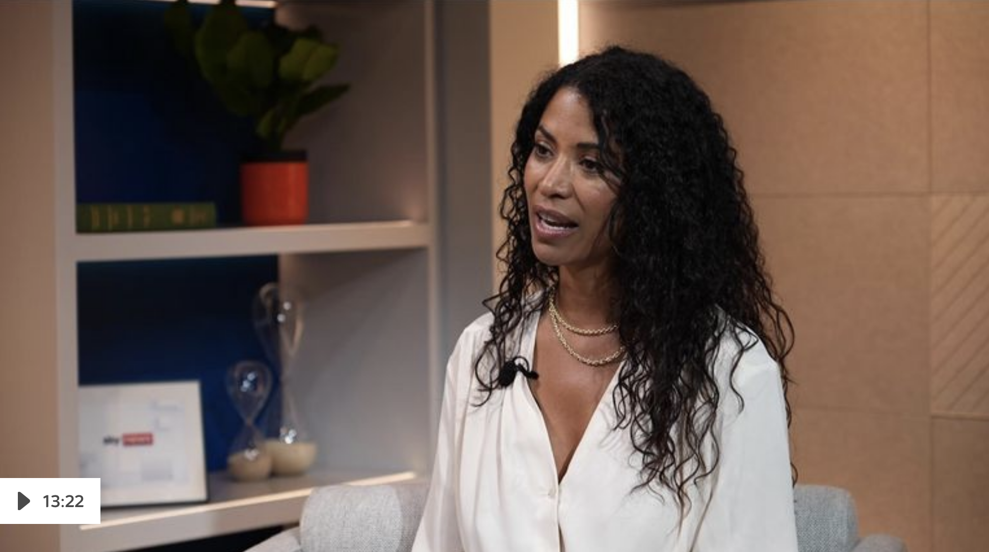 A woman with long, dark, curly hair wearing a white blouse and layered necklaces, speaking in a conversation space with a patterned wooden wall and a bookshelf with decorative items in the background.