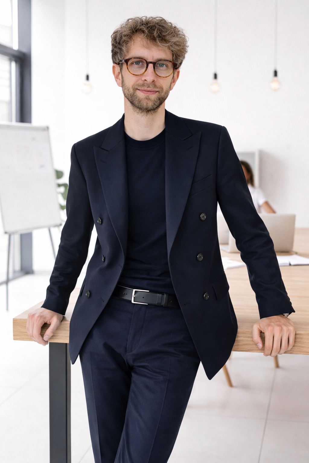 Uomo con capelli ricci e occhiali, vestito elegante in giacca e pantaloni blu, sorride in un ufficio moderno con illuminazione soffusa.
