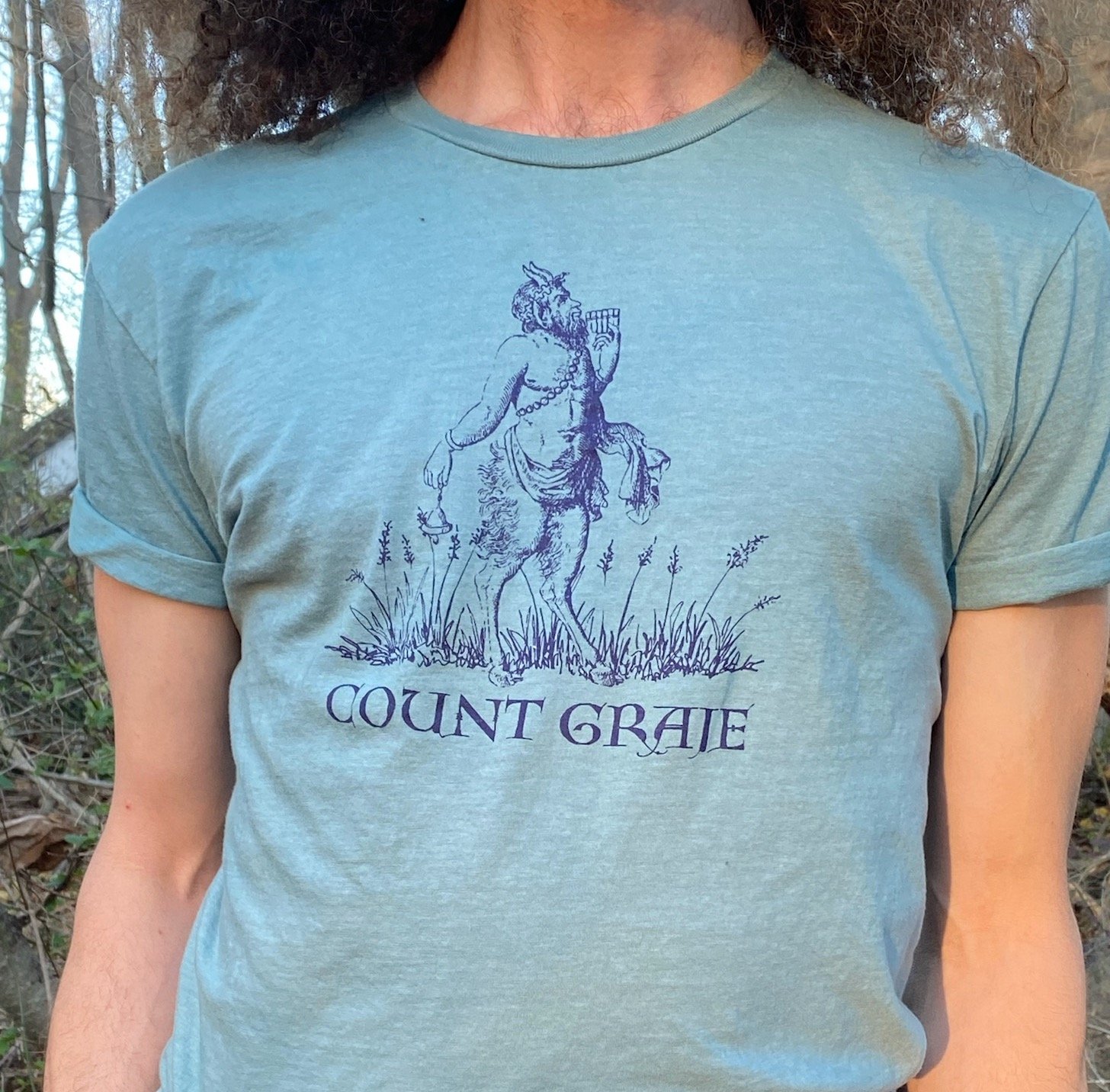 Count Graje T-Shirt a blue shirt with purple font and image of the Greek God Pan ringing a bell in the grass