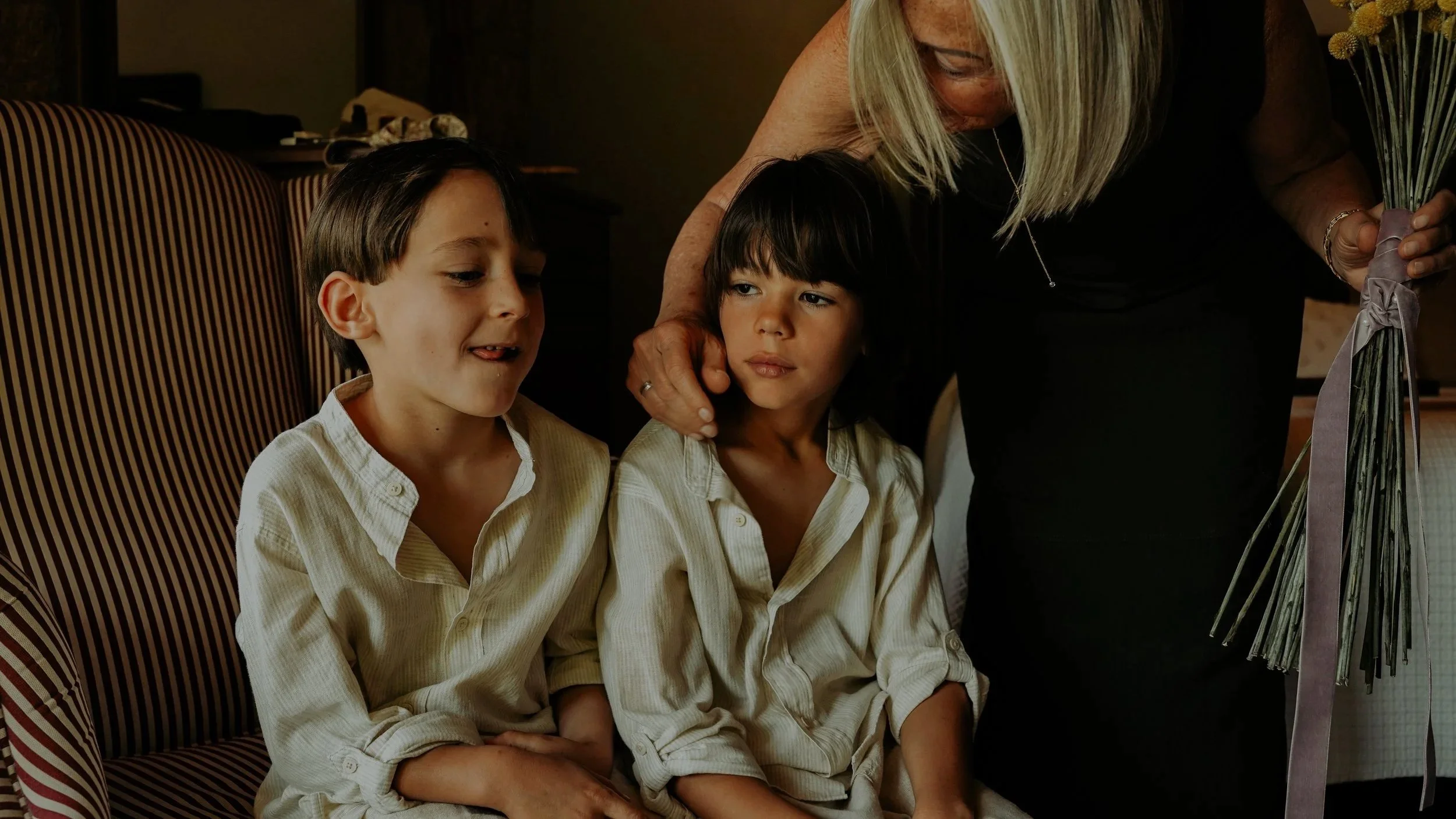 Dos niños con camisetas blancas sentados en un sillón y una mujer de cabello rubio y gafas, que sostiene un ramo de flores.