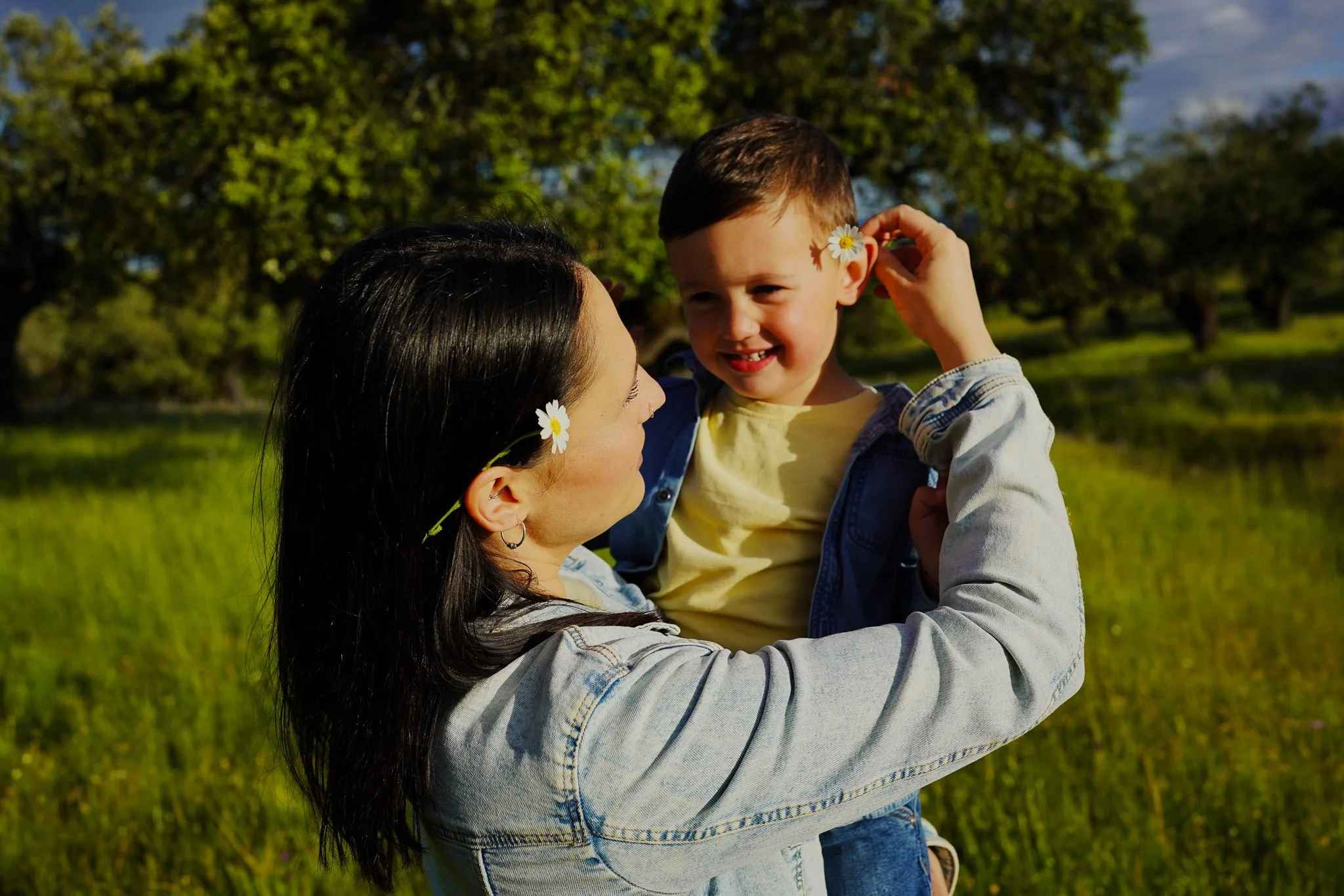 Una mujer y un niño con flores, mostrando un momento alegre en un campo verde con árboles y cielo azul.