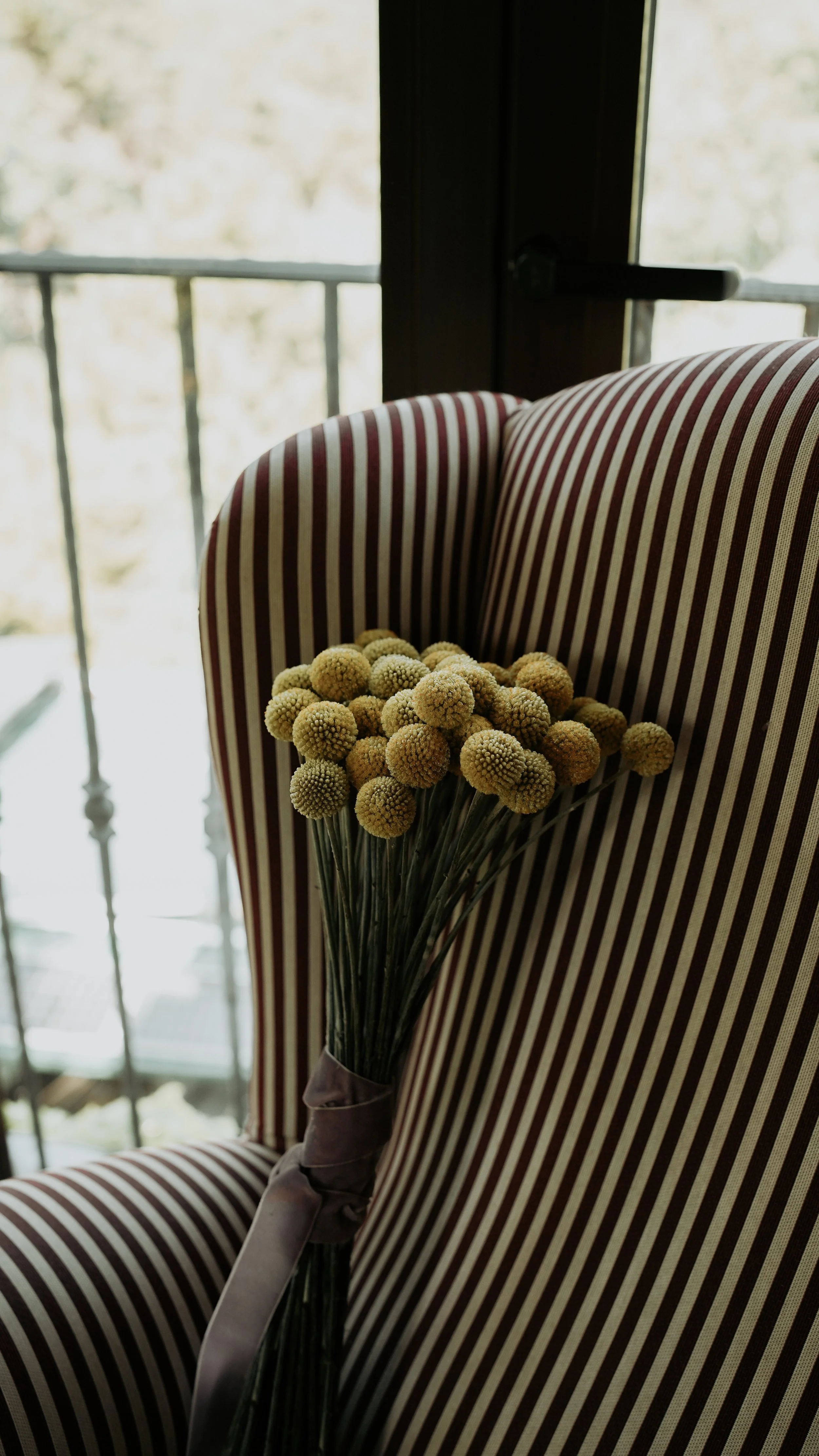 Ramo de flores amarillas sobre sillón de rayas en tonos beige y rojo en un interior con balcón y ventanas con luz natural.