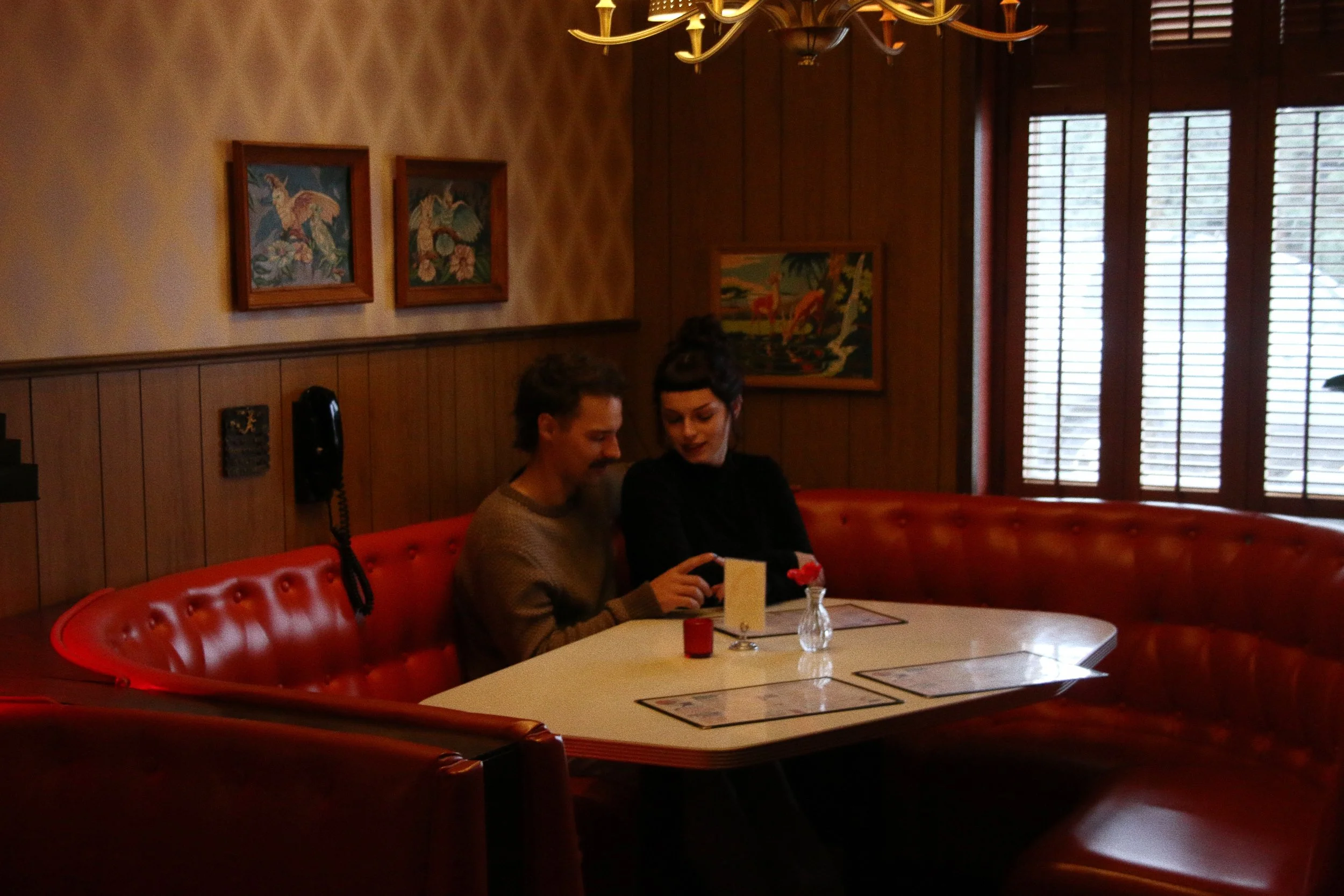 A man and a woman sitting together in a corner booth of a restaurant, looking at a smartphone, with a small flower vase and a napkin holder on the table.