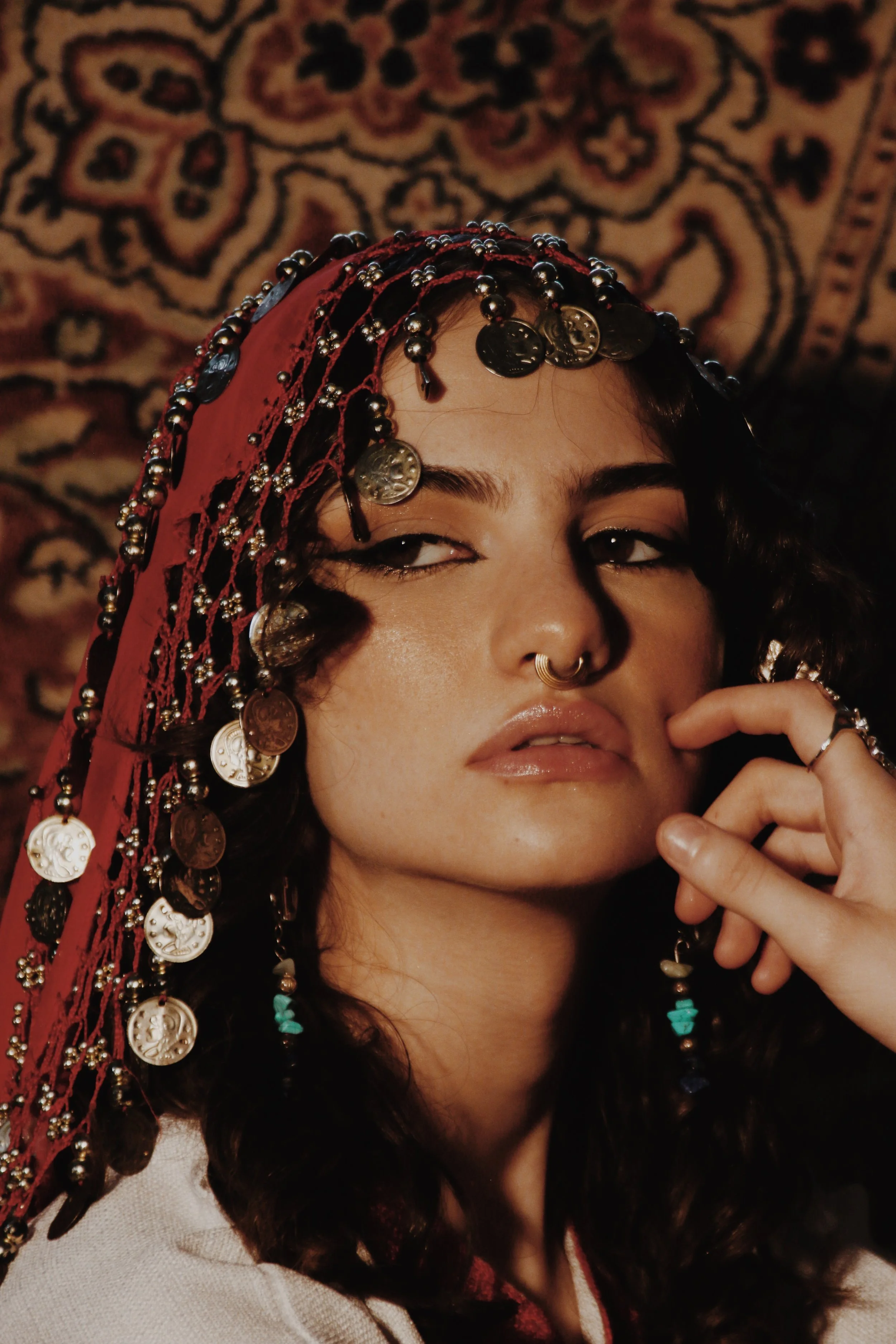 A woman with dark, curly hair wearing a headscarf decorated with coins and beads. She has a septum piercing, glossy lips, and is touching her face with her right hand. The background features a patterned rug.