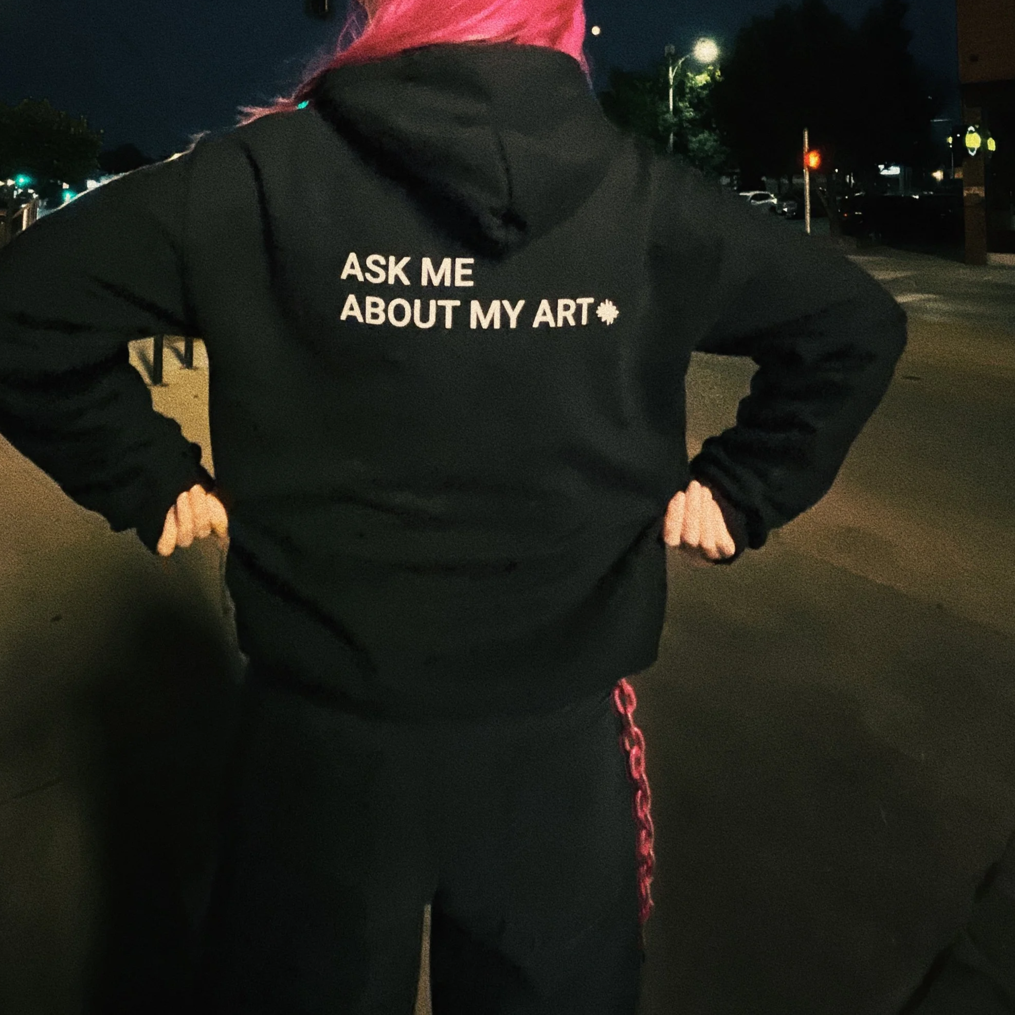Person with pink hair wearing a black hoodie that says 'Ask me about my art' on the back, standing outdoors at night.