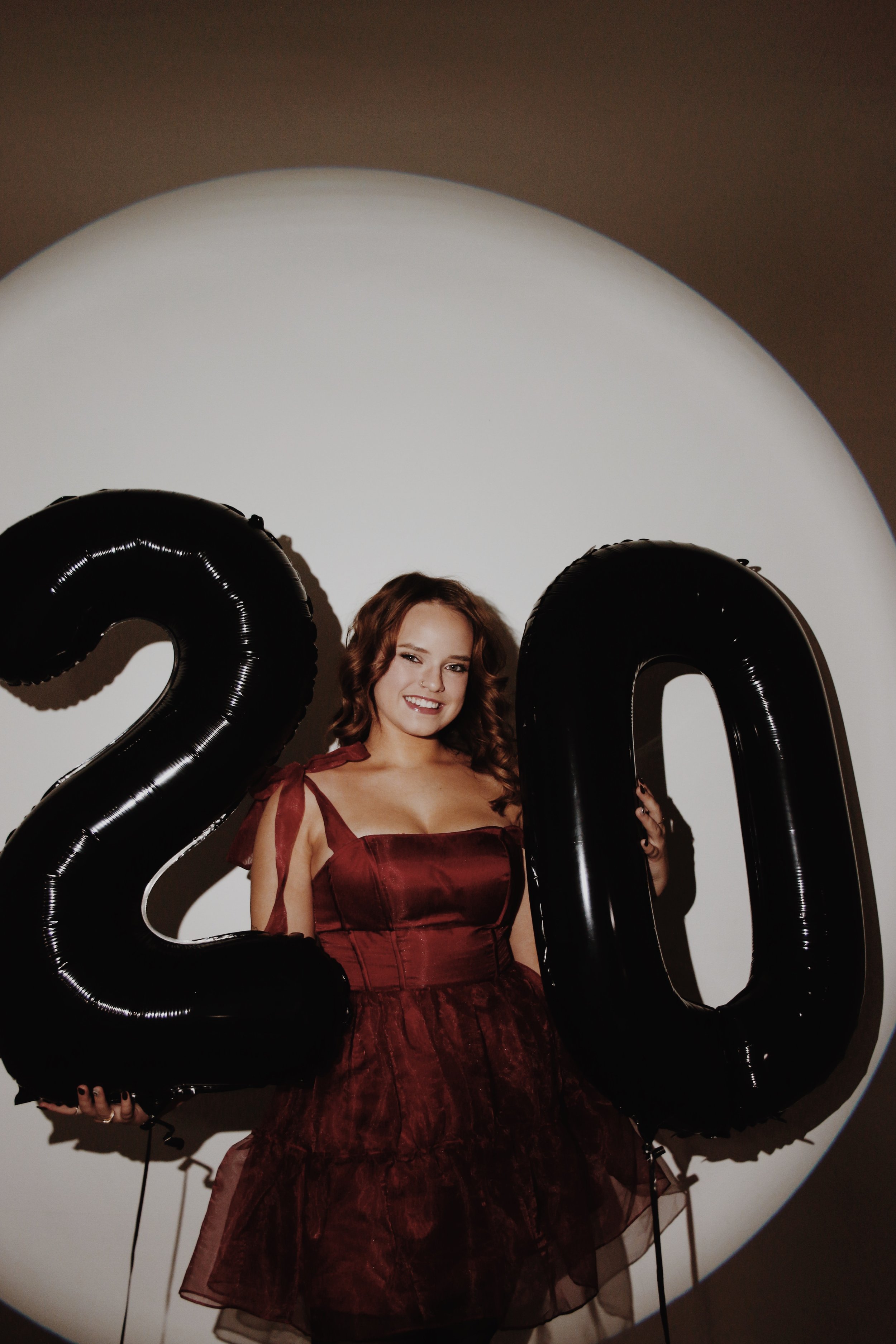 Young woman in burgundy dress holding black number balloons '2' and '0', standing against a white circular background.