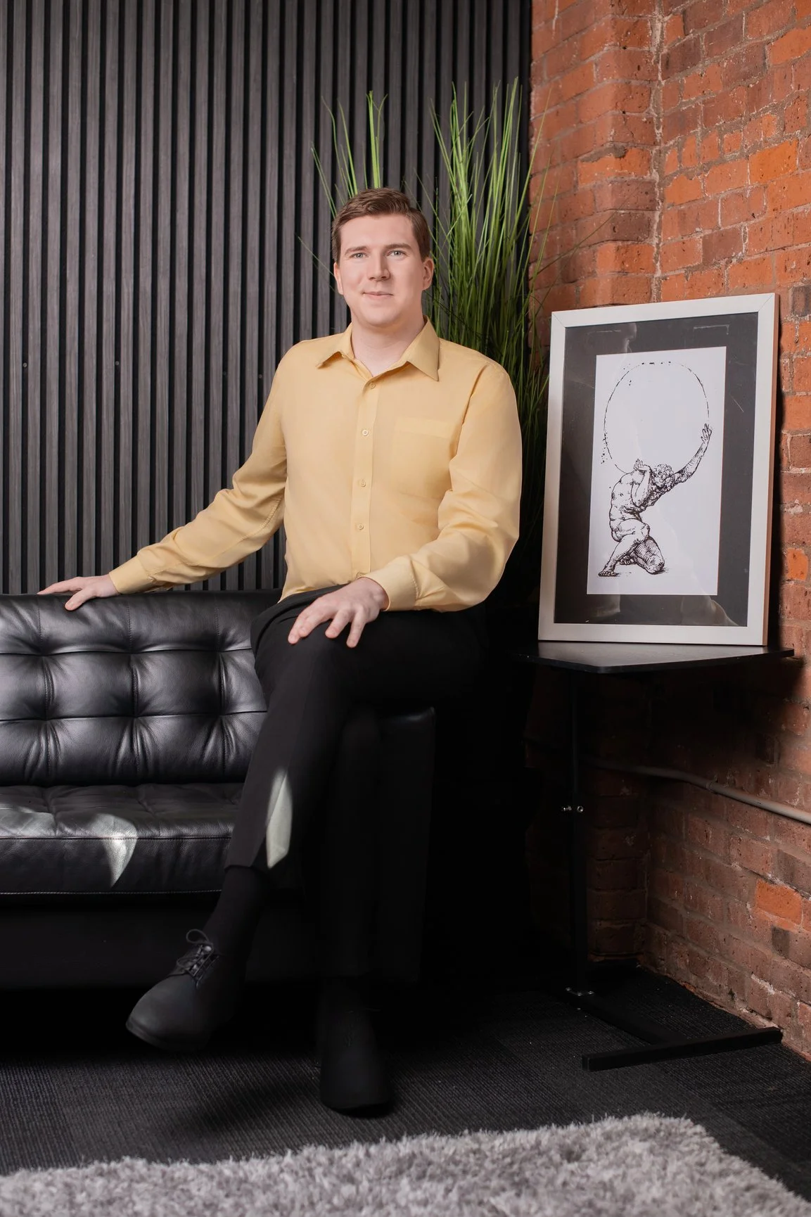 A man in a yellow shirt and black pants sitting on a black leather couch in a modern room with brick walls, black paneled wall, a tall green plant, and a framed sketch of a person balancing a circle on their head.