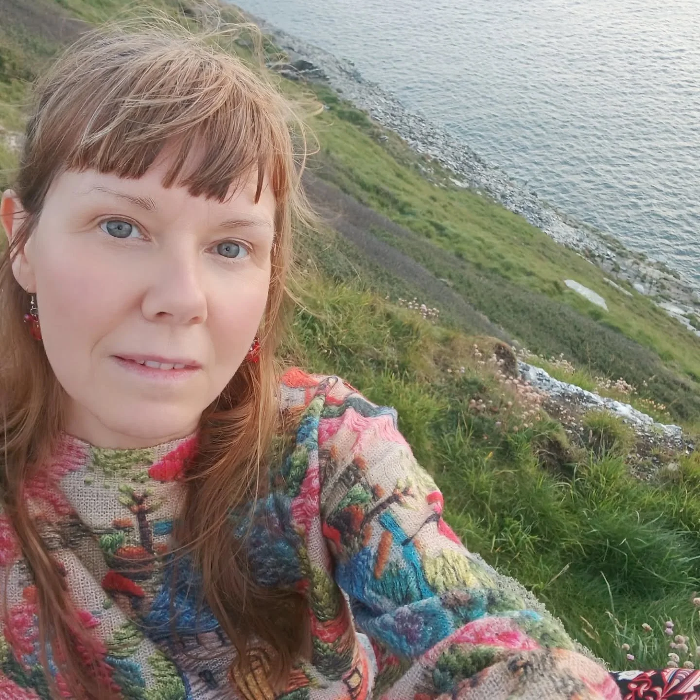 Selfie of a woman with red hair, blue eyes, wearing a colorful patterned top, standing on a grassy hillside near a body of water.