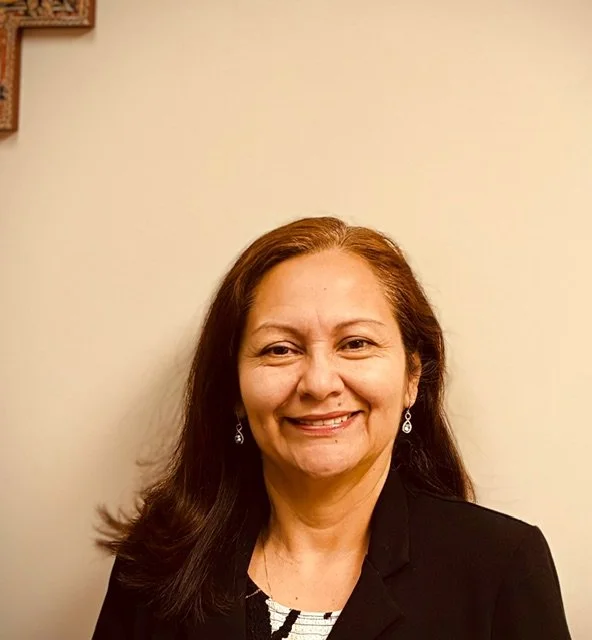 Rina Richers who is on the Board of Directors for RHC with shoulder-length dark hair smiling, wearing earrings and a black blazer, standing against a plain light-colored wall.