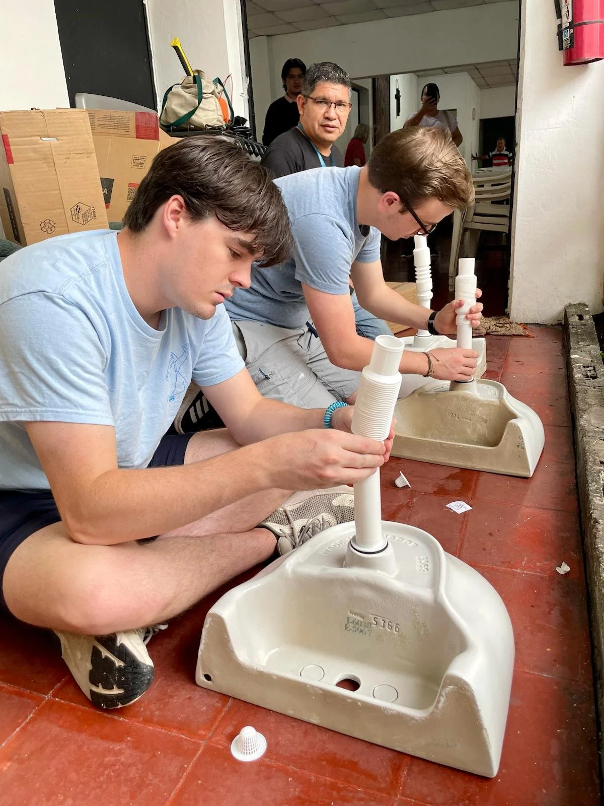 Two young men sitting on the floor in front of sinks, working on assembling or repairing plumbing fixtures. Behind them, an older man looks at the camera, and other people are visible in the background.