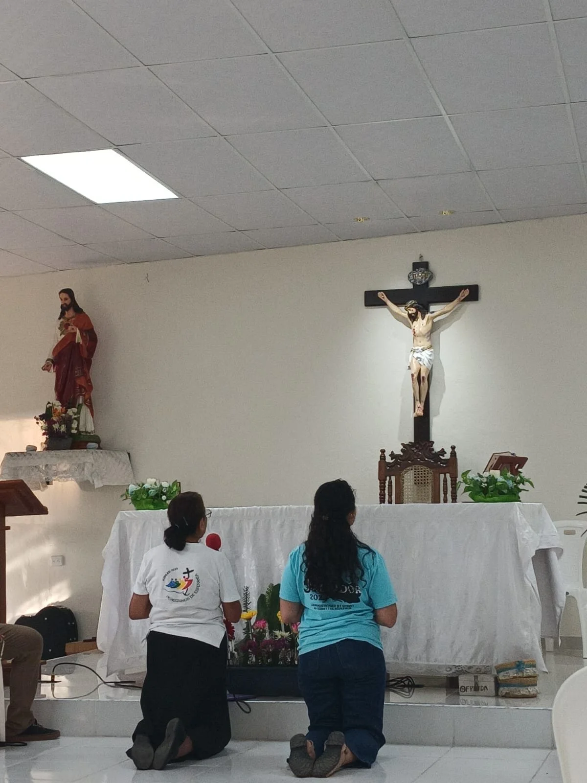 Two women kneel in prayer before an altar with a crucifix and statue of Jesus Christ, inside the Milagroso Chapel.