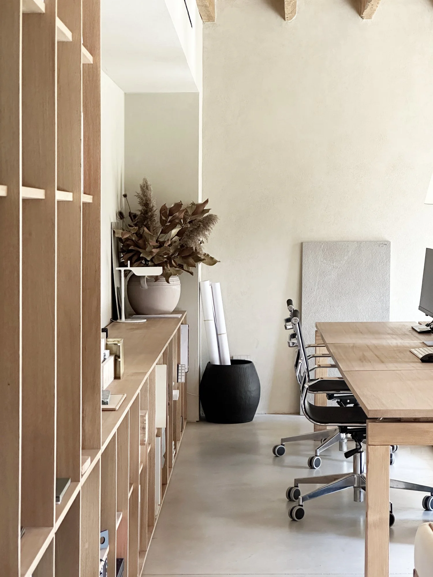 Interno di un ufficio minimalista con mobili in legno chiaro, una grande sala con tavolo e sedie ergonomiche, un grande vaso nero con piante essiccate e una scrivania con oggetti.