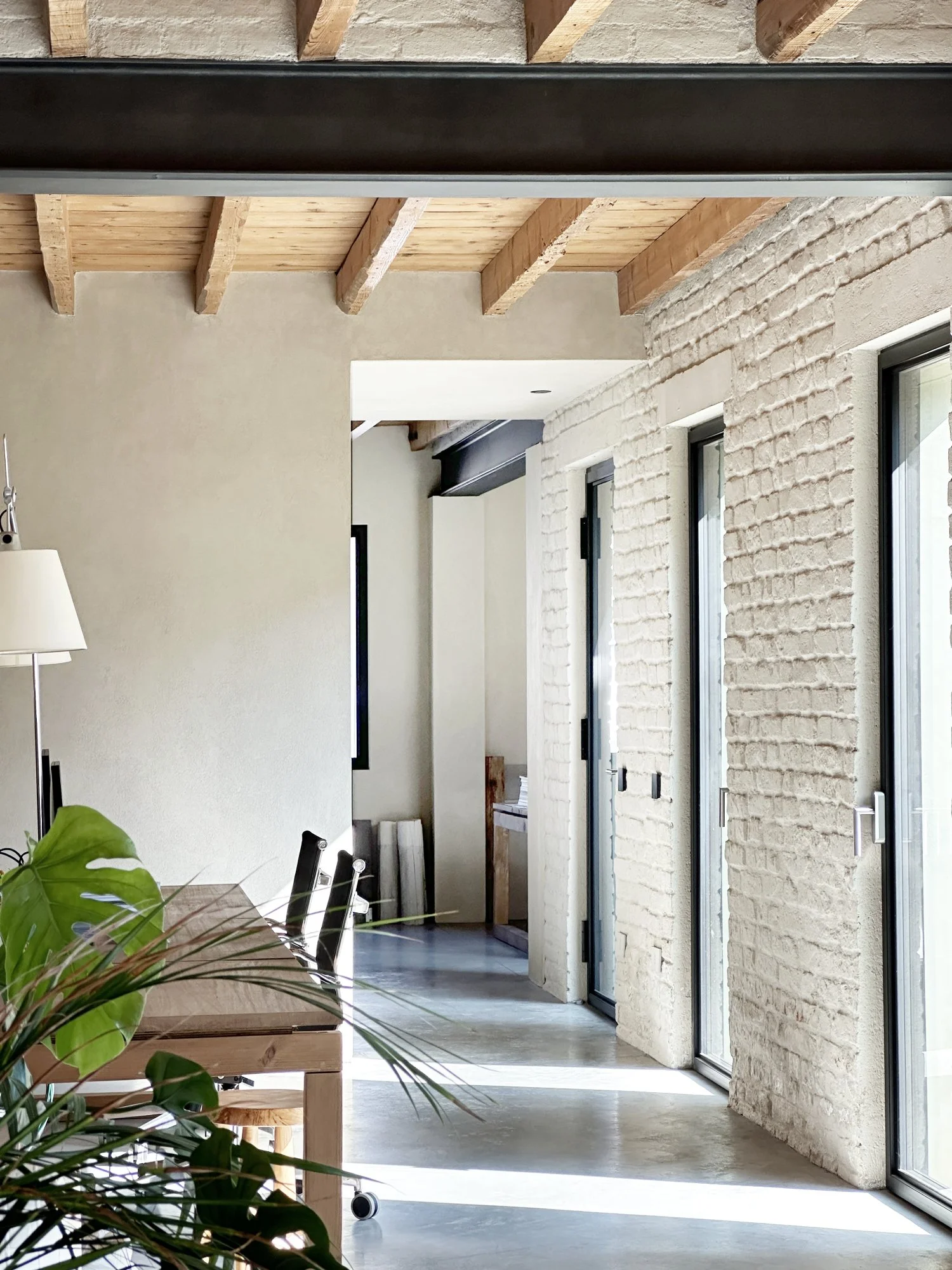Interno di una stanza luminosa con pareti in mattoni bianca, grandi porte in vetro e soffitto con travi di legno.