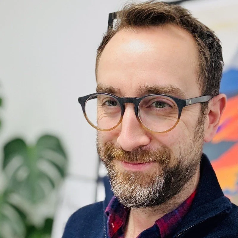 Close-up of a smiling man with glasses, beard, and mustache, wearing a dark jacket and a red plaid shirt, indoors with a blurred colorful background and a plant visible.