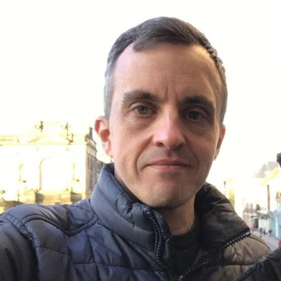 A man with short dark hair and blue eyes taking a selfie outdoors, wearing a dark quilted jacket, with sunlight illuminating the background buildings.