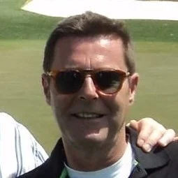 Man with short dark hair wearing sunglasses outdoors, smiling, with a green grassy background.