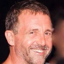 A smiling man with short brown hair and a beard, wearing a dark shirt, with a dark background